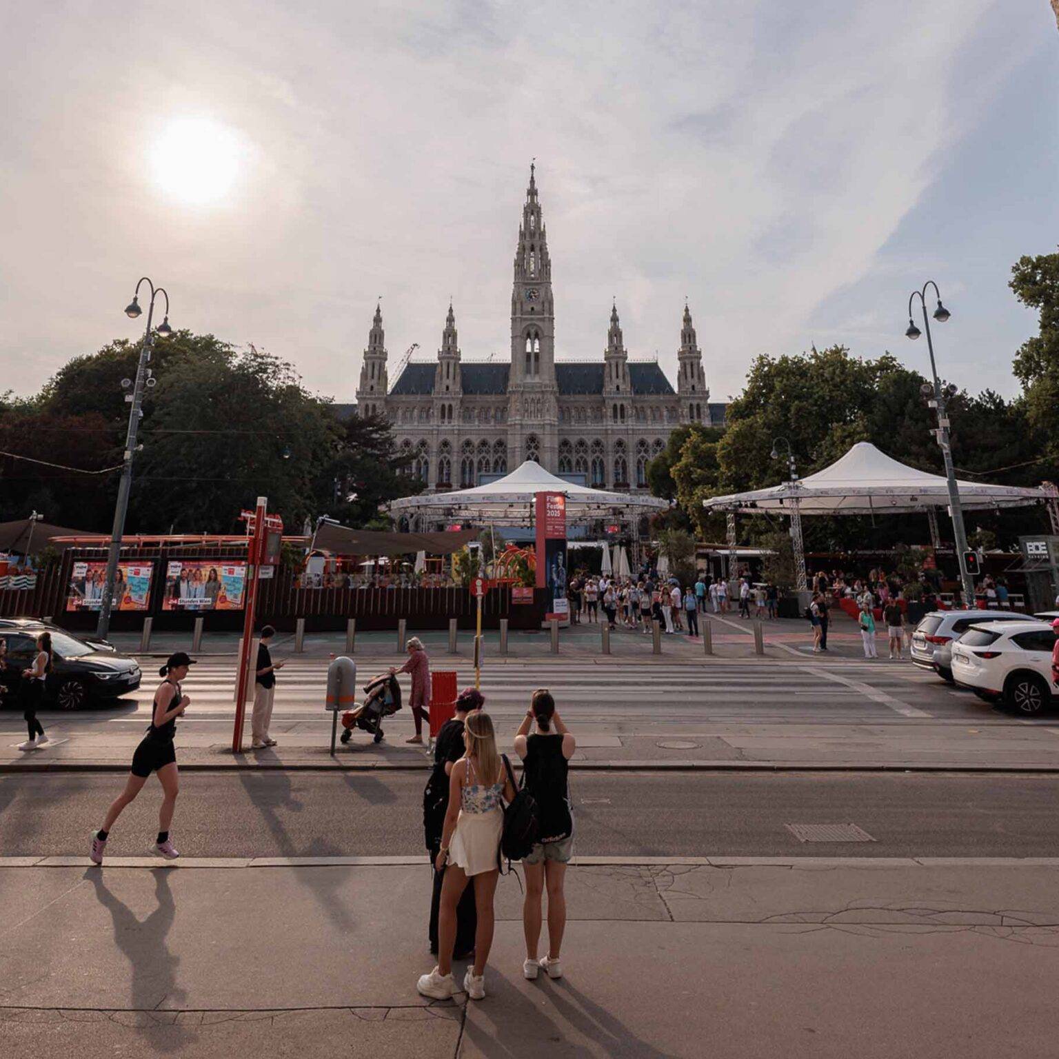 Blick auf das Rathaus von Wien, ein großes Gebäude mit Turm, umgeben von Menschen und Autos, bei Sonnenlicht.