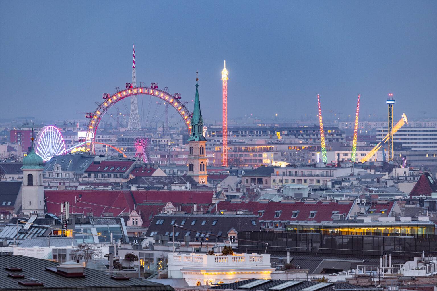 Die Skyline von Wien und der Prater mit bunten Lichtern und einem Riesenrad bei Dämmerung.