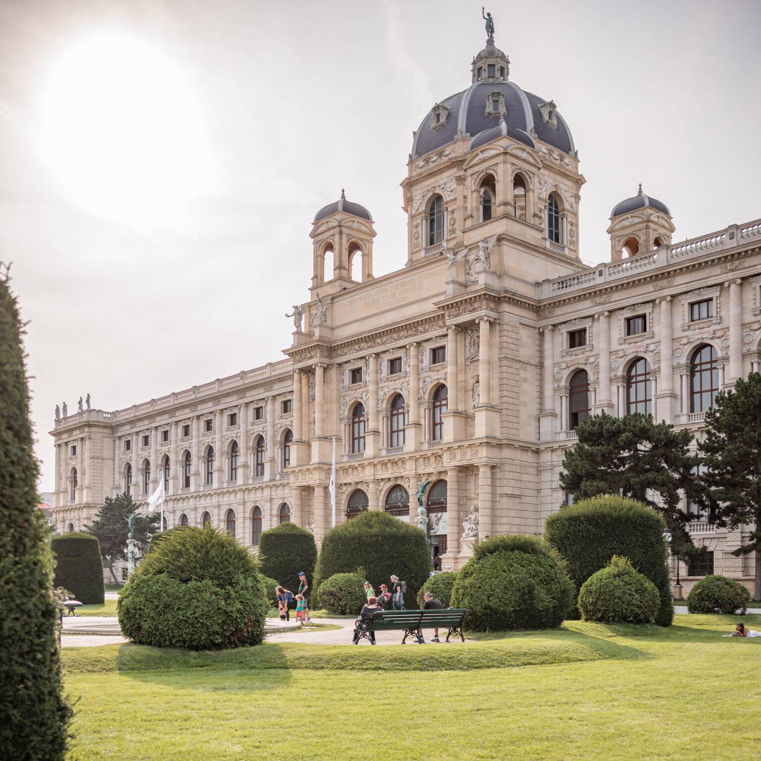Historische Fassade des NHM mit Kuppel, umgeben von gepflegten Gärten und Besuchern im Freien.