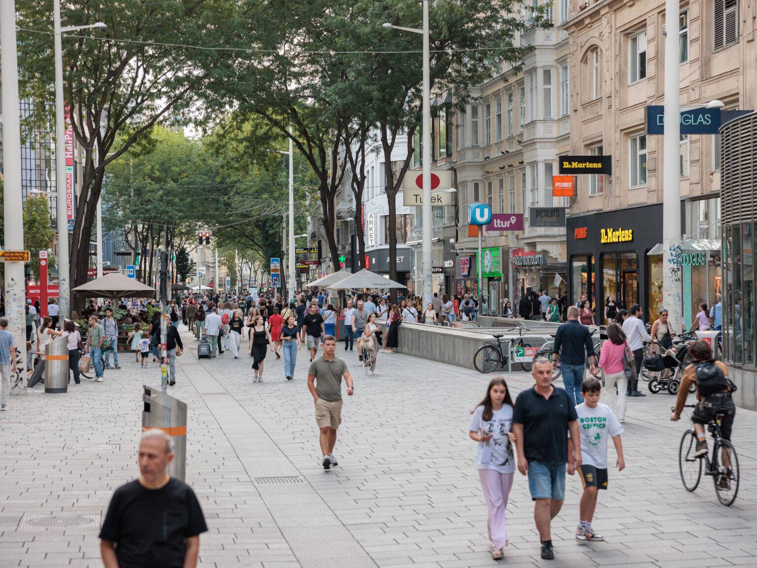 Belebte Einkaufsstraße Mariahilfer mit Fußgängern, Bäumen und Geschäften, darunter Cafés und Boutiquen.