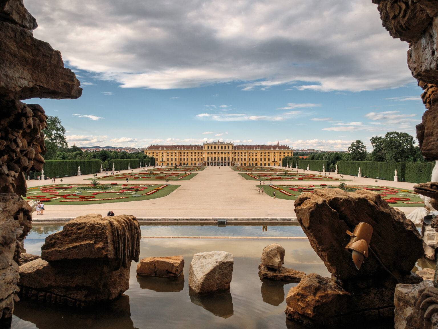 Blick durch Felsen auf das Schloss Schönbrunn und die Gartenanlagen mit Blumenbeeten und Wasserflächen.