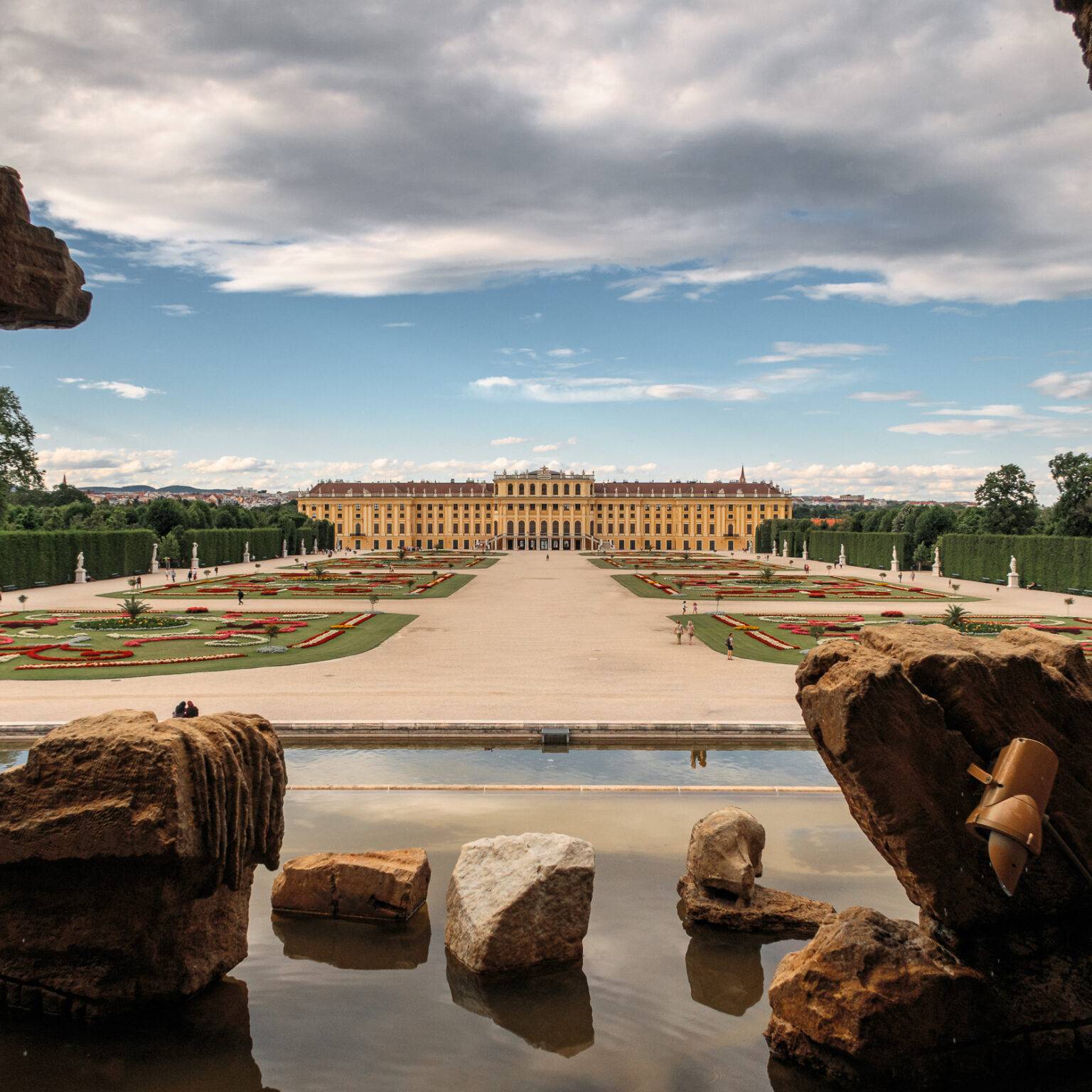 Blick durch Felsen auf das Schloss Schönbrunn und die Gartenanlagen mit Blumenbeeten und Wasserflächen.