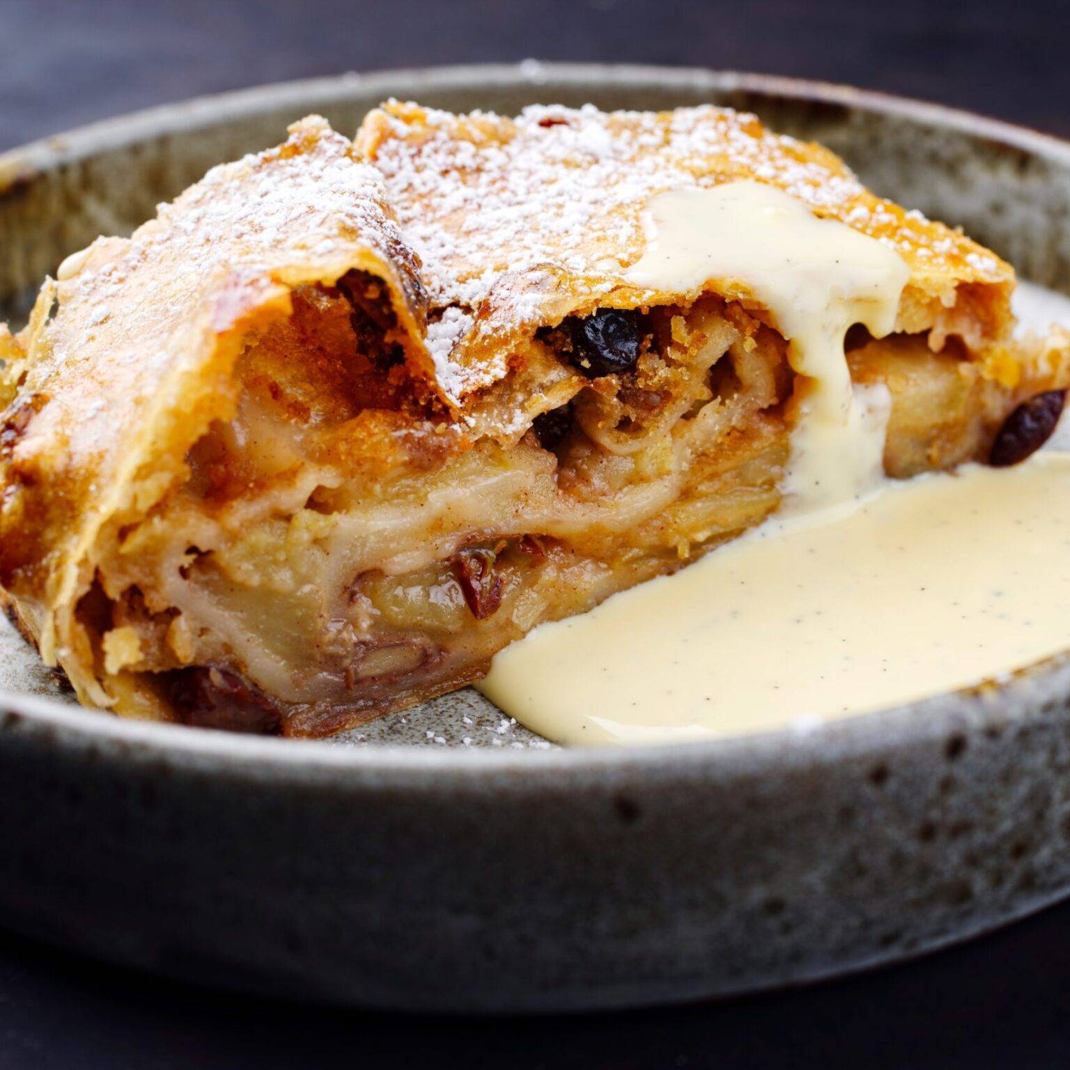 Ein Stück Apfelstrudel mit Puderzucker und einer Portion Vanillesoße auf einem grauen Teller.