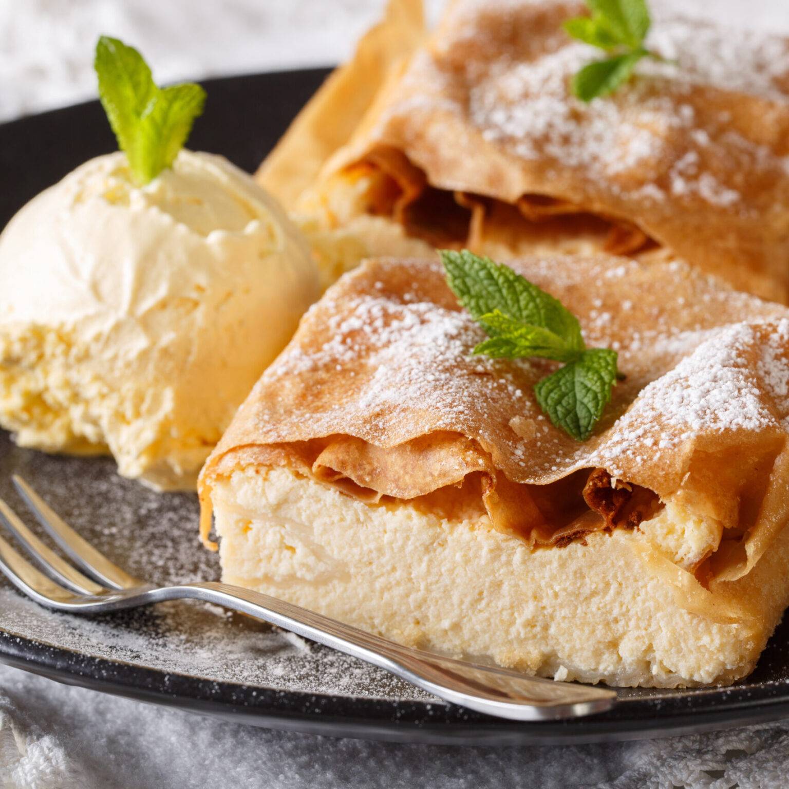 Zwei Stücke Topfenstrudel, Blätterteig mit Quarkfüllung, mit Puderzucker und Minze, serviert mit einer Kugel Vanilleeis auf einem schwarzen Teller.