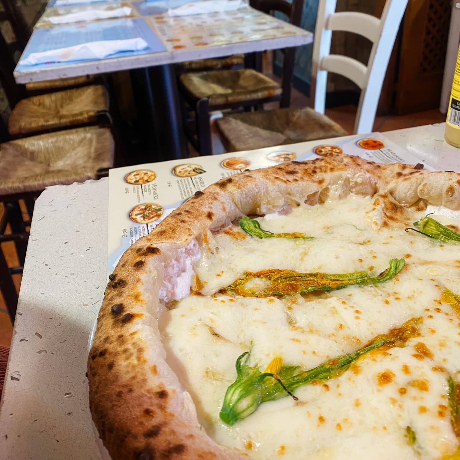 Eine große Pizza auf einem Teller, dahinter typische rattanbezogene Stühle.