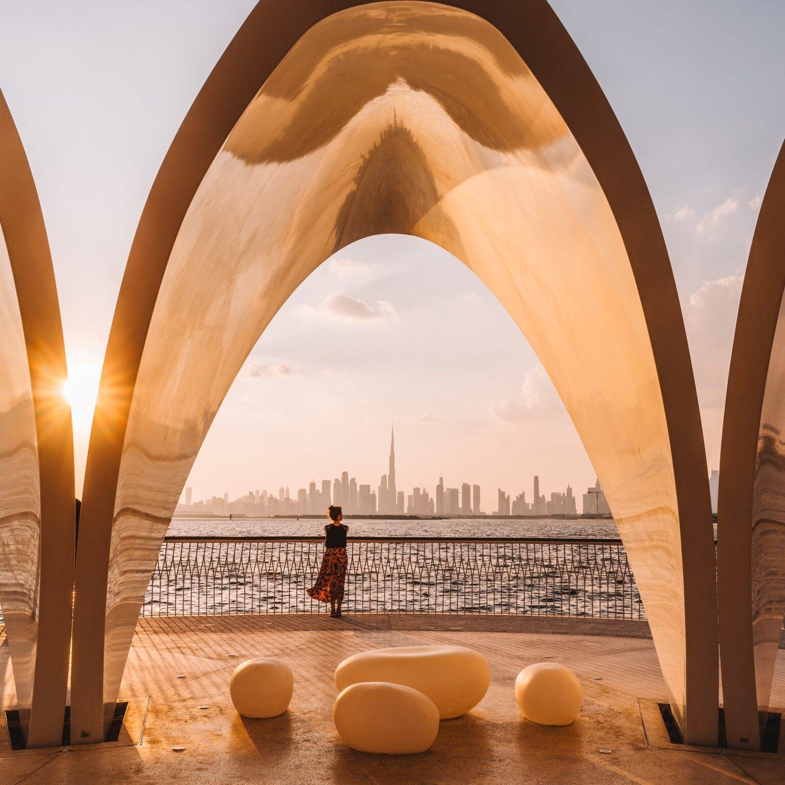 Ein goldenes ovales Tor. Dahinter steht eine Frau am Geländer zum Creek Habour in Dubai mit vielen Hochhäusern in Hintergrund.
