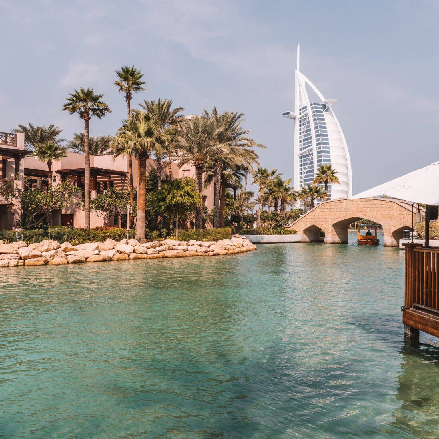 Ein kleiner Fluss mit Gebäuden und Palmen links und rechts und dem Burj al Arab Hotel im Hintergrund.