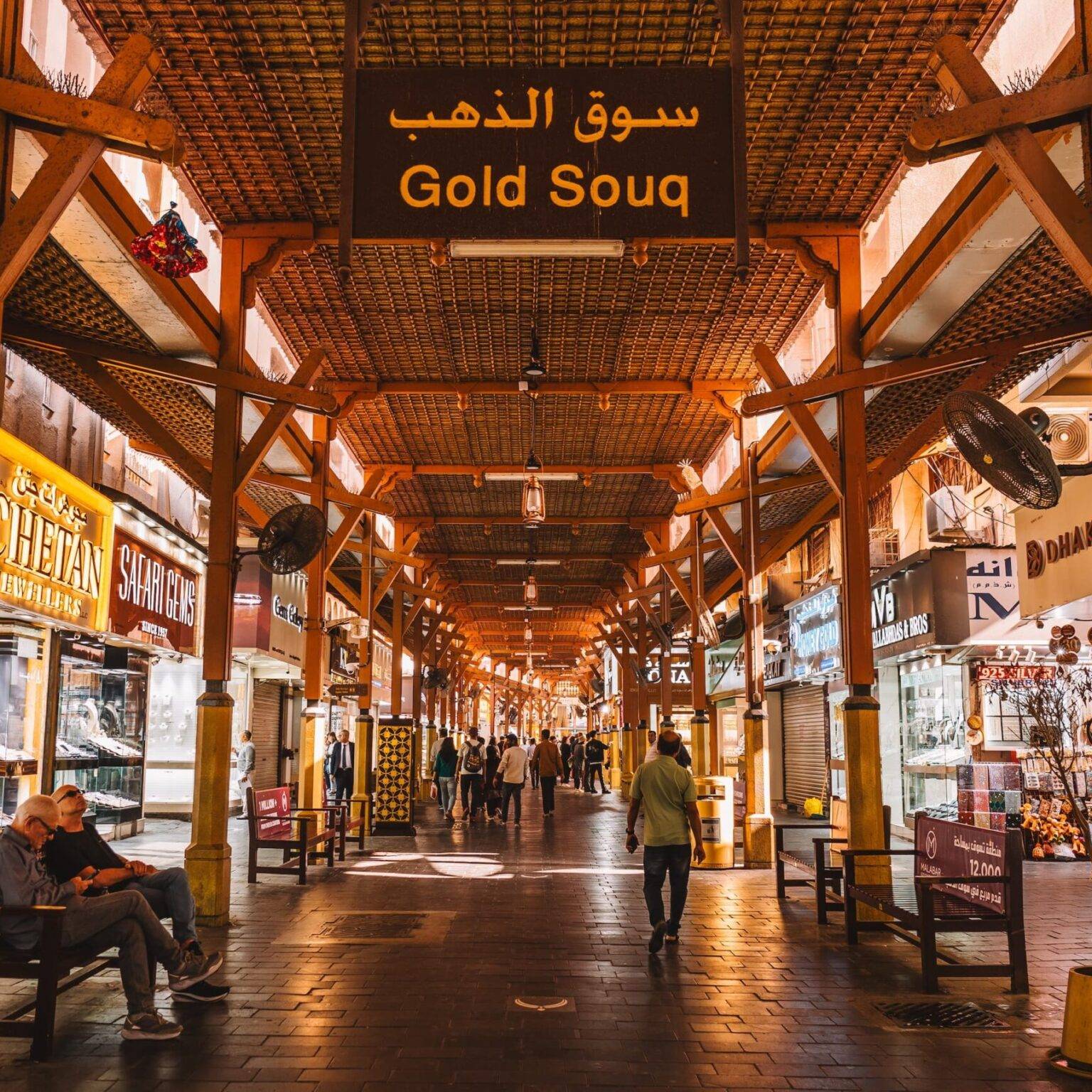 Ein Schild mit der Aufschrift "Gold Souq" und dahinter eine überdachter Gehweg gesäumt von Schmuckgeschäften links und rechts.