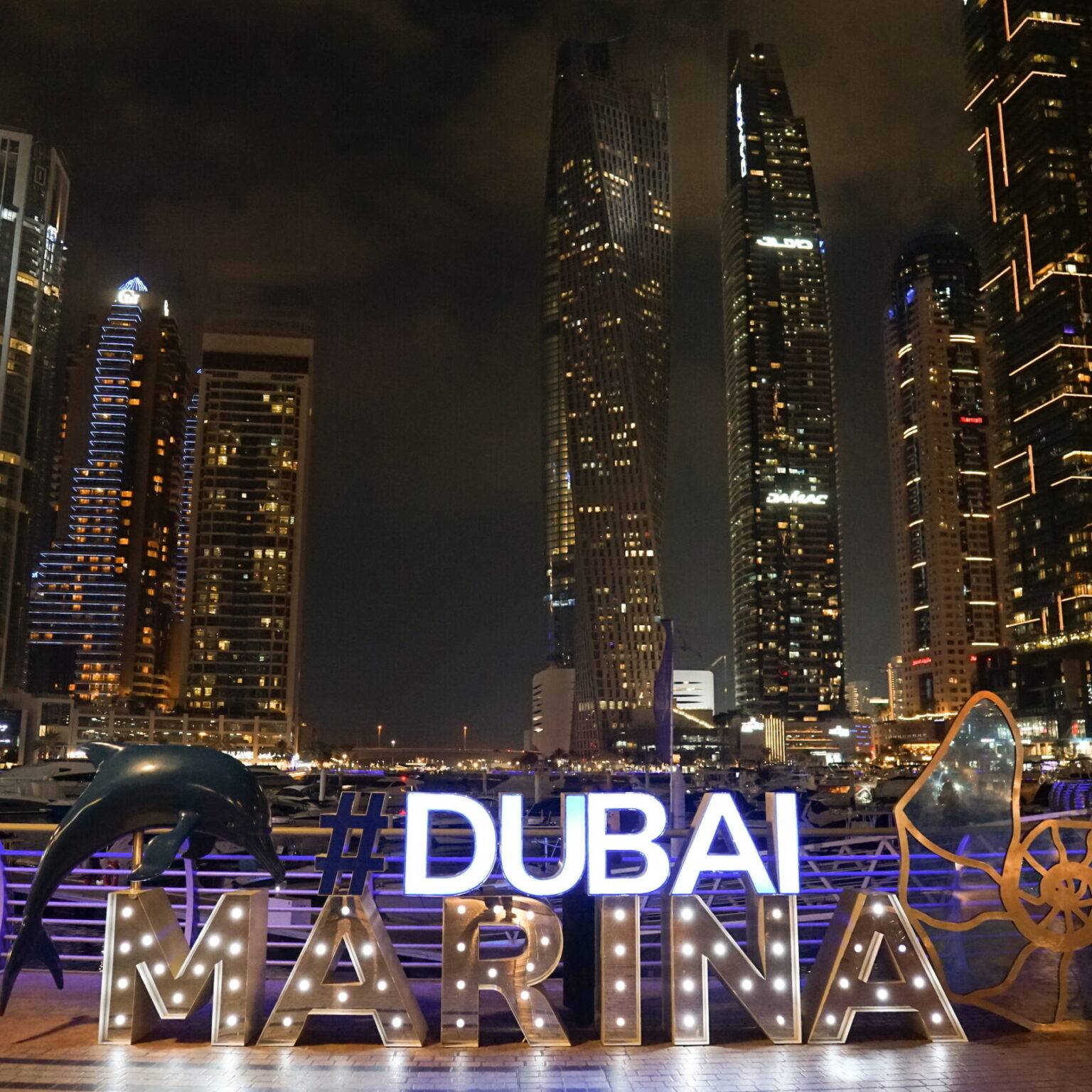 Beleuchtete Buchstaben bilden den Schriftzug "#DUBAI MARINA". Im Hintergrund sind beleuchtete Hochhäuser bei Nacht.