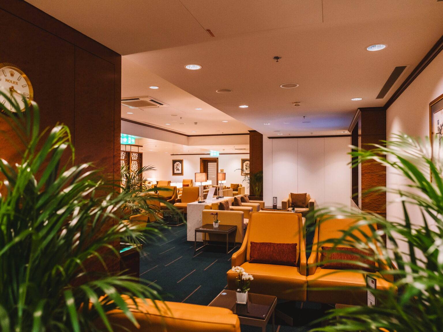 Der Sitzbereich der Emirates Lounge am Flughafen München mit orangefarbenen Ledersesseln, dunkelblauem Teppichboden, dunklen Holzdekorationen und einigen Pflanzen im Vordergrund.
