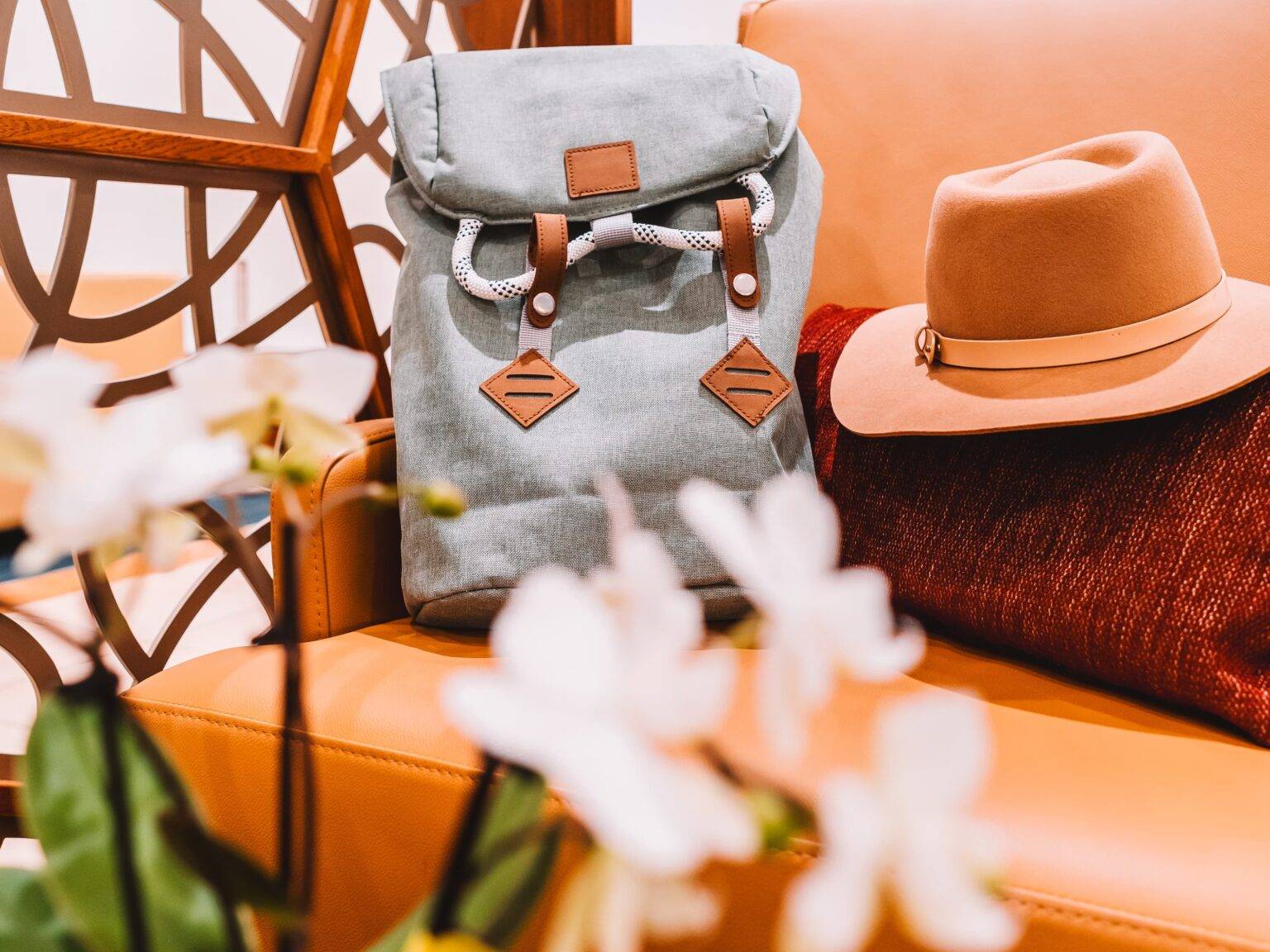 Auf einer orangefarbenen Ledercouch liegt ein Rucksack und ein Hut. Im Vordergrund steht eine Orchidee.