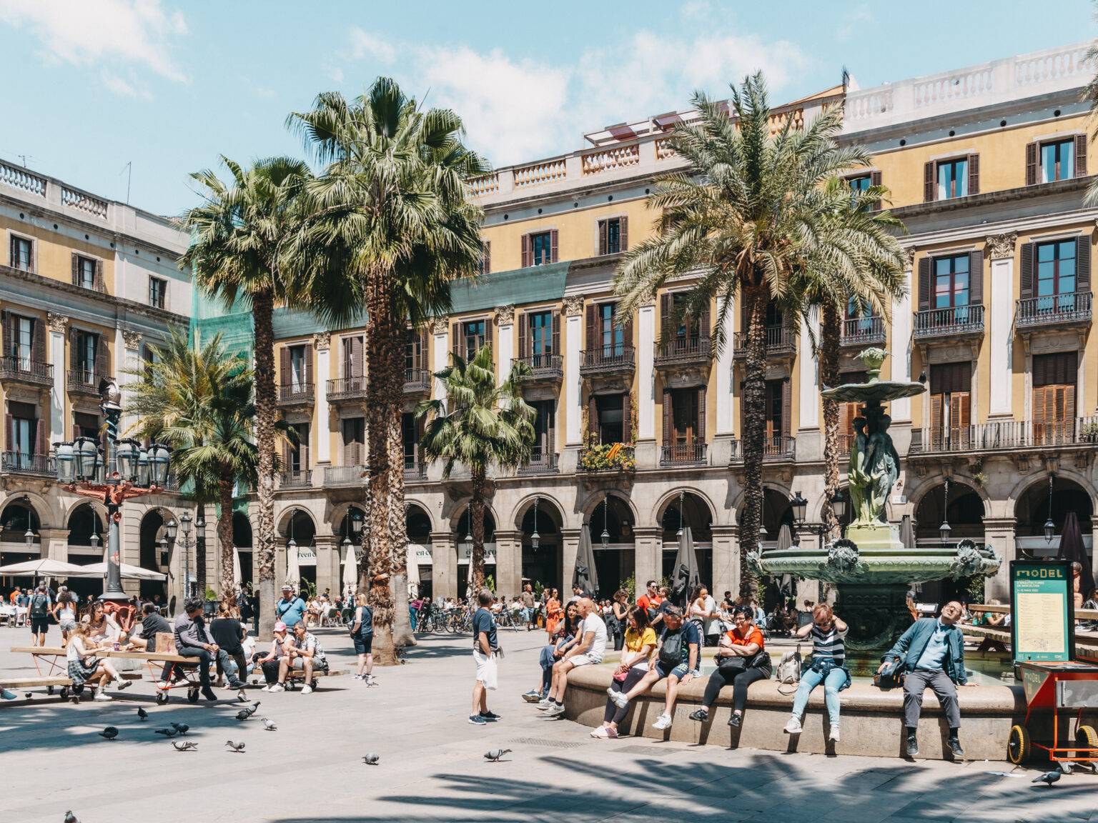 Der Plaça Reial, ein Platz mit einem Brunnen und mehreren Palmen, umgeben von Wohnhäusern. Der Platz ist voller Passanten.