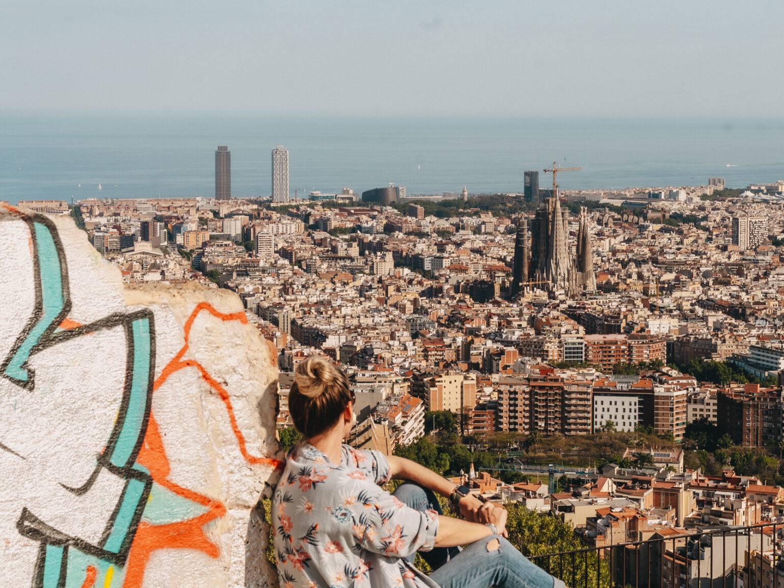 Die Bloggerin Nina sitzt auf der mit Graffiti besprühten Ruine des Bunkers del Carmel und blickt über Barcelona. Die Türme der Sagrada Familia ragen heraus. Im Hintergrund das Meer.