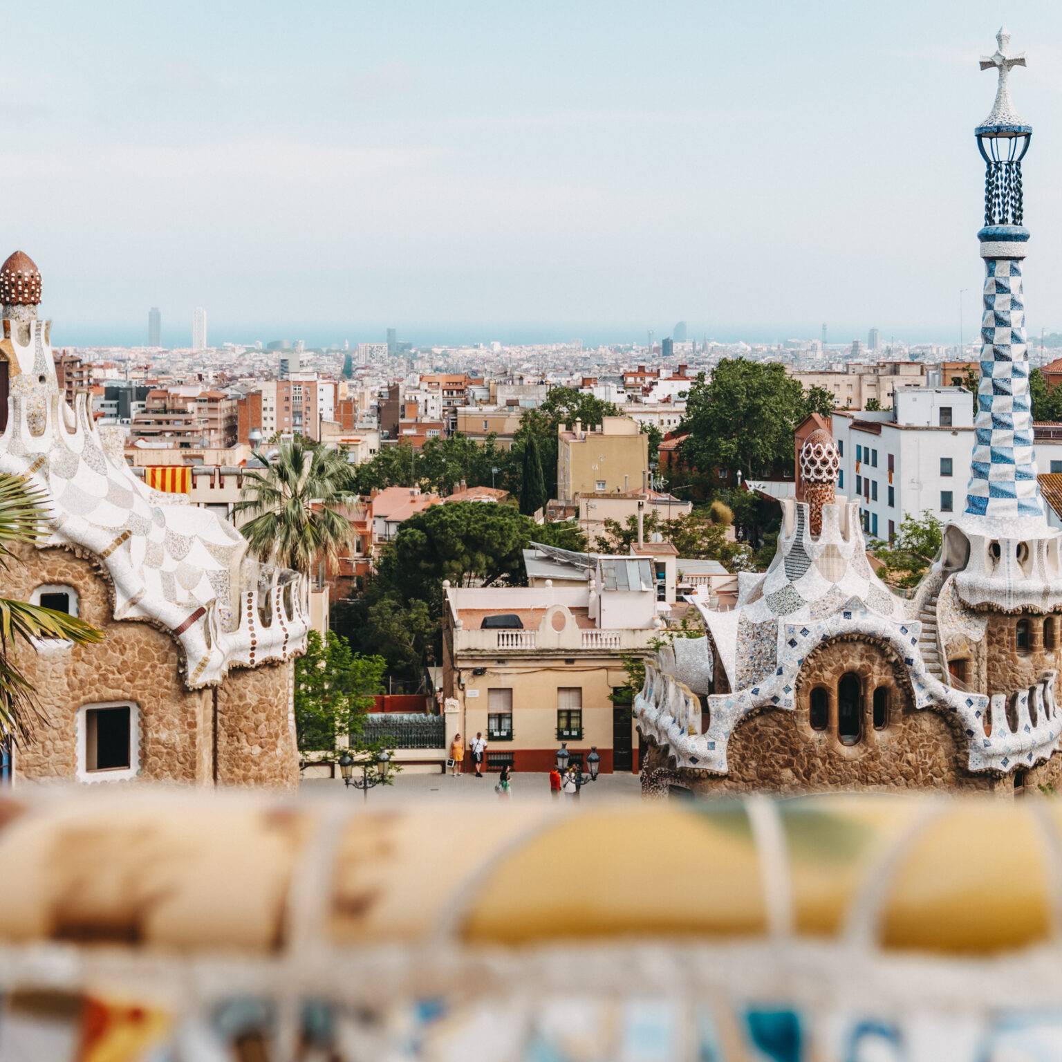 Die Skyline von Barcelona aus der Perspektive des Park Guell. Im Vordergrund sind die mit Mosaik verzierten Gebäude des Eingangs des Parks und im Hintergrund das Meer.
