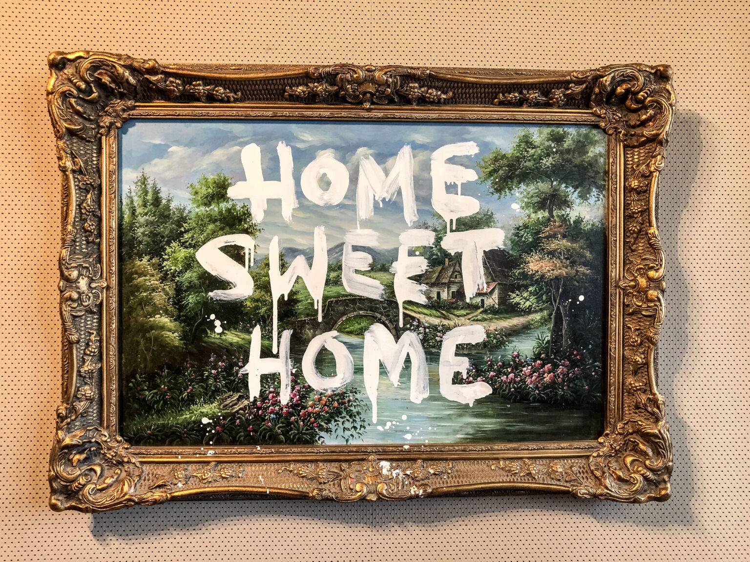 Ein Kunstwerk von Banksy mit Landschaftsmotiv. Darüber wurde mit weißer Farbe "HOME SWEET HOME" geschrieben.