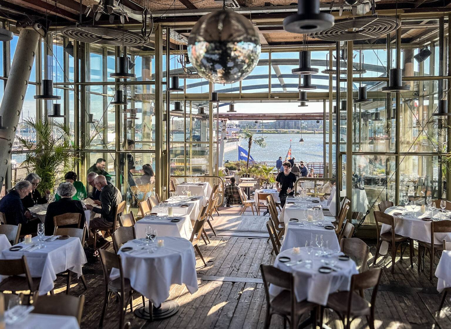 Innenansicht des Restaurants Helling 7 in Amsterdam-Noord mit stilvollem Gastraum und Panoramablick auf das Wasser durch große Fenster.