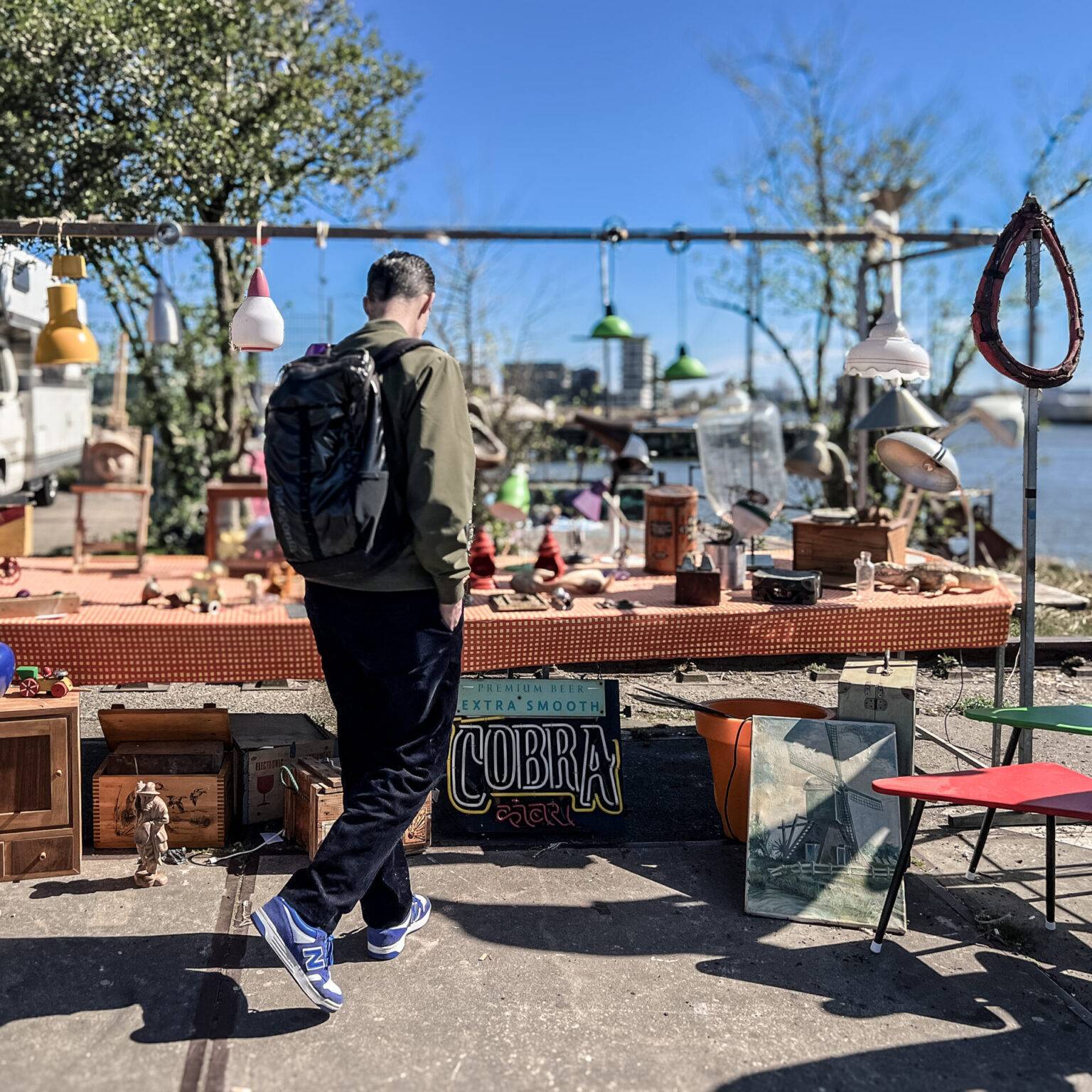 Mann mit Rucksack betrachtet einen Flohmarktstand mit Vintage-Artikeln und bunten Lampen am Wasser in Amsterdam-Noord bei sonnigem Wetter.