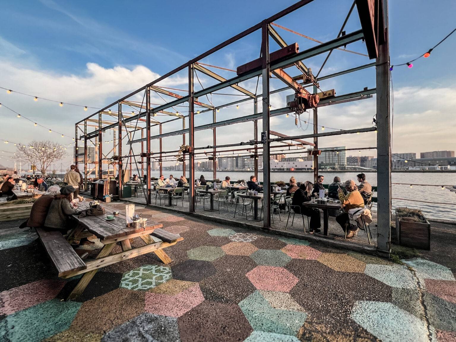 Menschen sitzen auf der Sonnenterrase des Restaurants Lowlander in Amsterdam Noord direkt am Wasser.