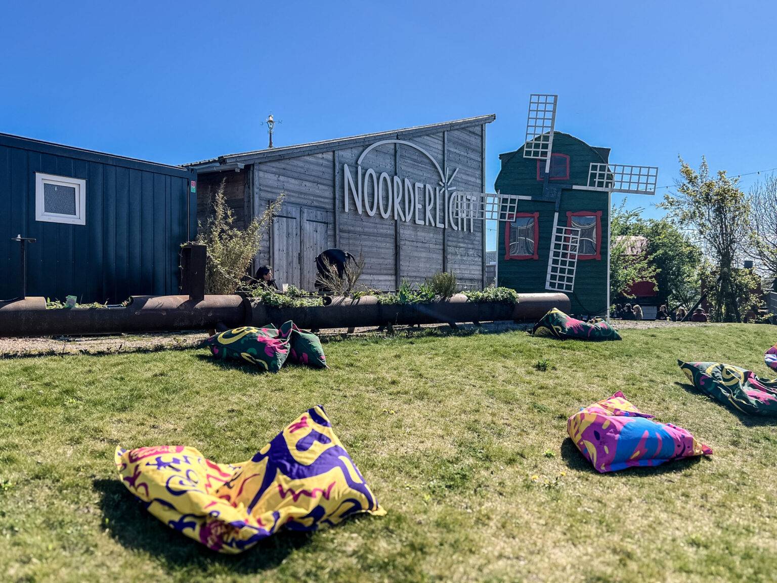 Außenbereich des Restaurants Noorderlicht in Amsterdam-Noord mit bunten Sitzsäcken auf der Wiese, sichtbarem Logo und einer dekorativen Holzmühle im Hintergrund.