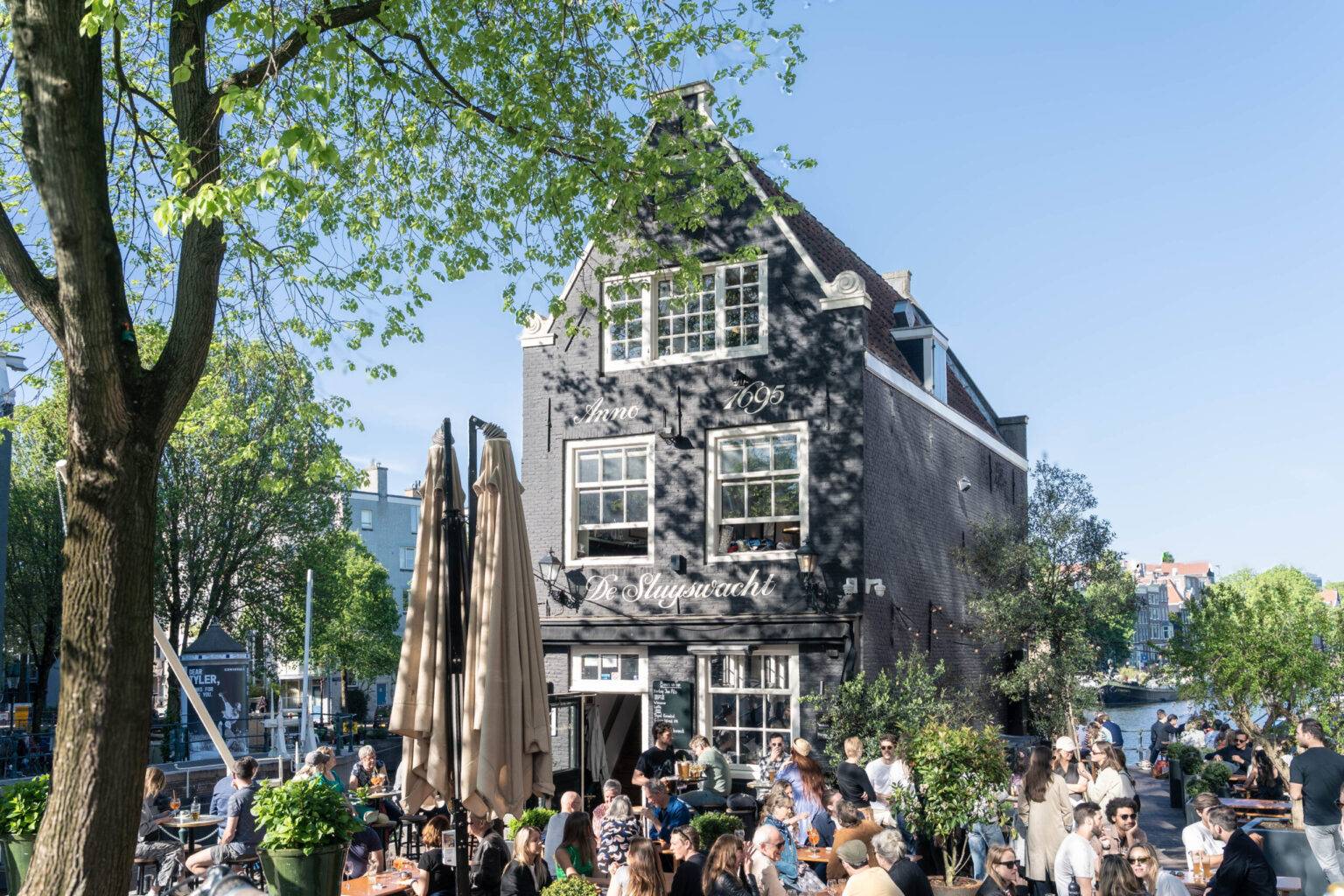 Menschen sitzen an einem sonnigen Tag vor dem Café de Sluyswacht in Amsterdam direkt an der Gracht und genießen die Atmosphäre.
