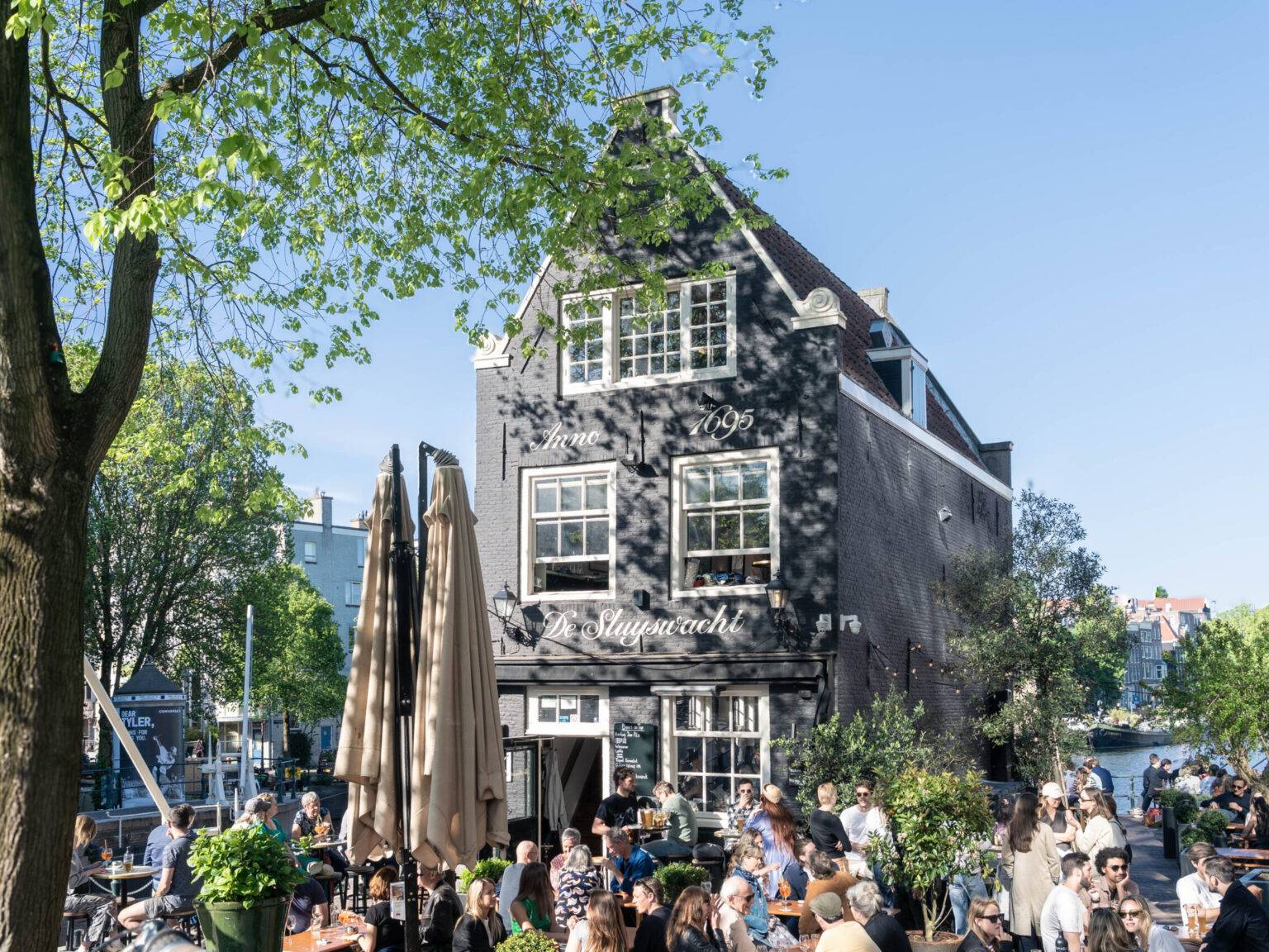Menschen sitzen an einem sonnigen Tag vor dem Café de Sluyswacht in Amsterdam direkt an der Gracht und genießen die Atmosphäre.