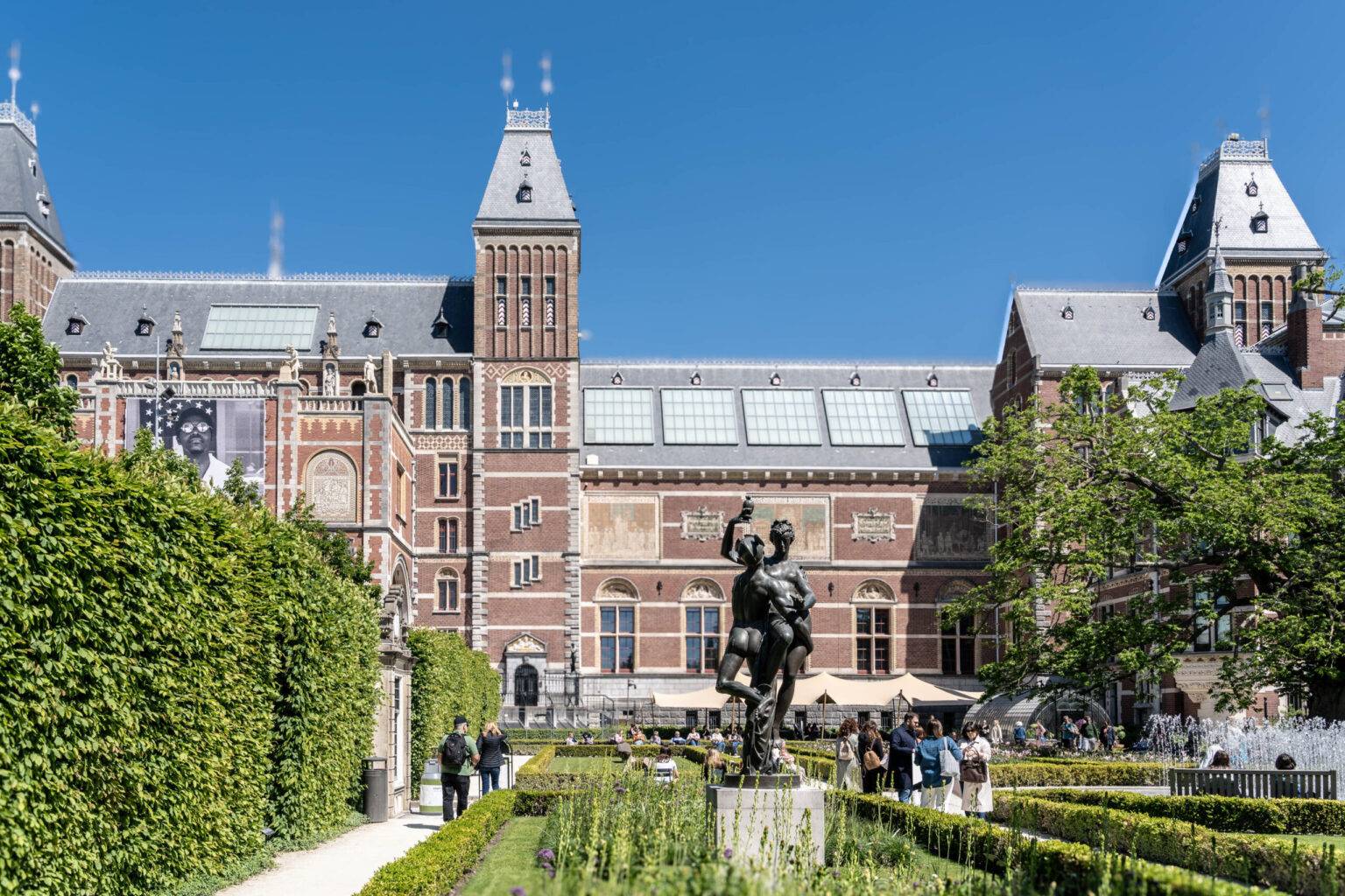 Das Rijksmuseum in Amsterdam mit seinem kunstvoll angelegten Garten bei Tageslicht, Besucher flanieren zwischen Blumenbeeten und Skulpturen.