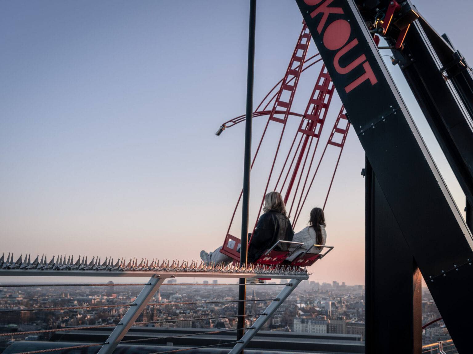 Zwei Personen schaukeln auf dem A’DAM Tower bei Sonnenuntergang mit weitem Blick über Amsterdam.