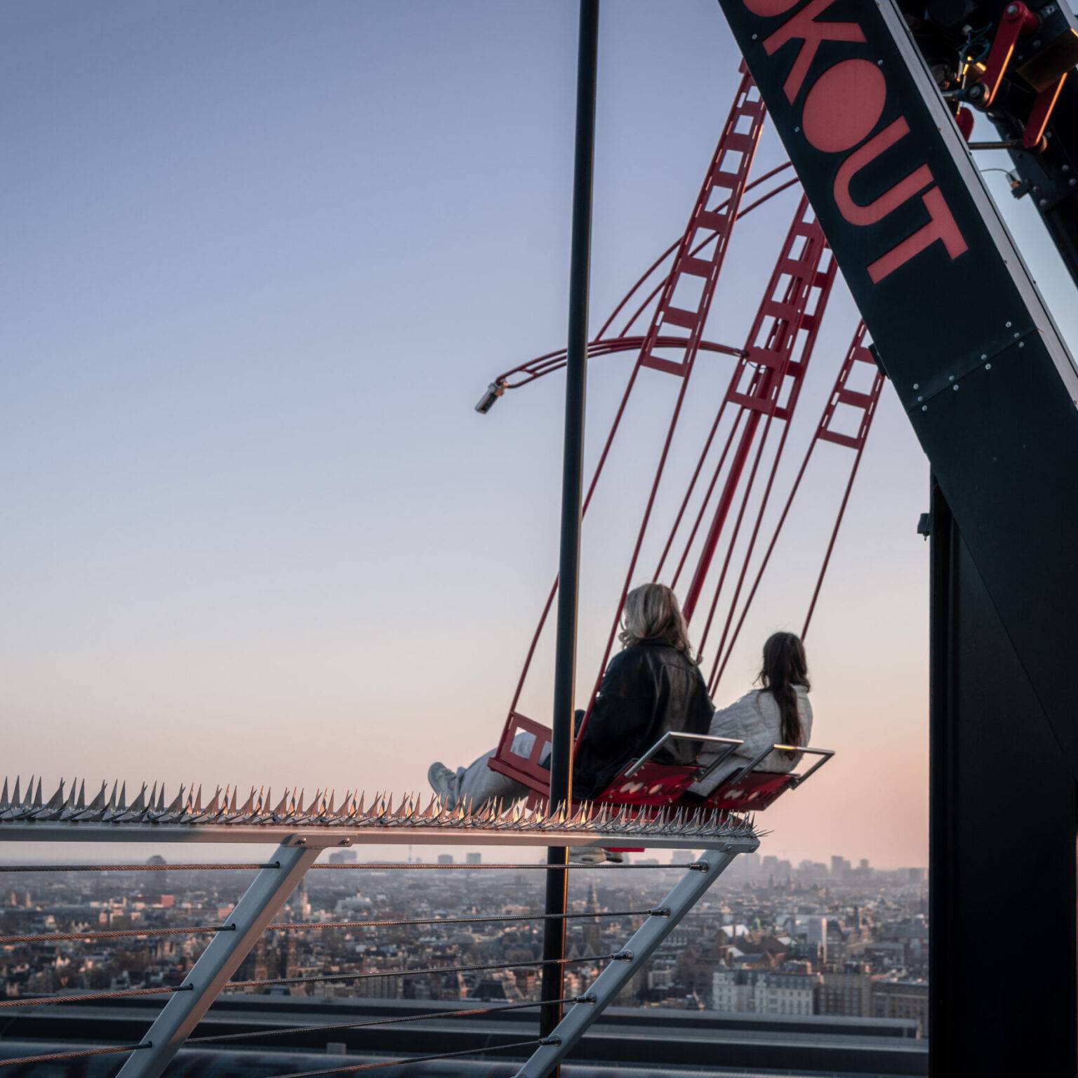 Zwei Personen schaukeln auf dem A’DAM Tower bei Sonnenuntergang mit weitem Blick über Amsterdam.