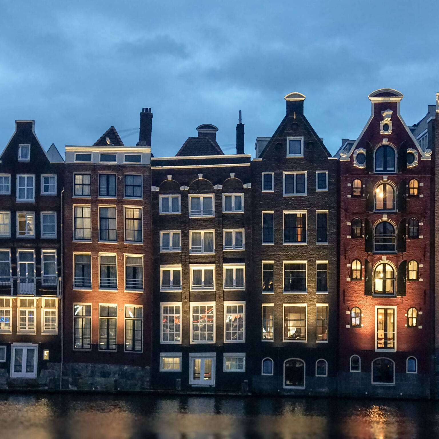Historische Giebelhäuser im beleuchteten Amsterdam Centrum am Wasser bei Dämmerung – beliebte, aber stark besuchte Touristenkulisse.