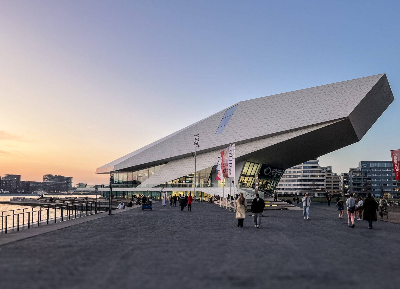 Das Eye Filmmuseum in Amsterdam-Noord bei Sonnenuntergang mit moderner, futuristischer Architektur und Spaziergängern entlang der Uferpromenade.