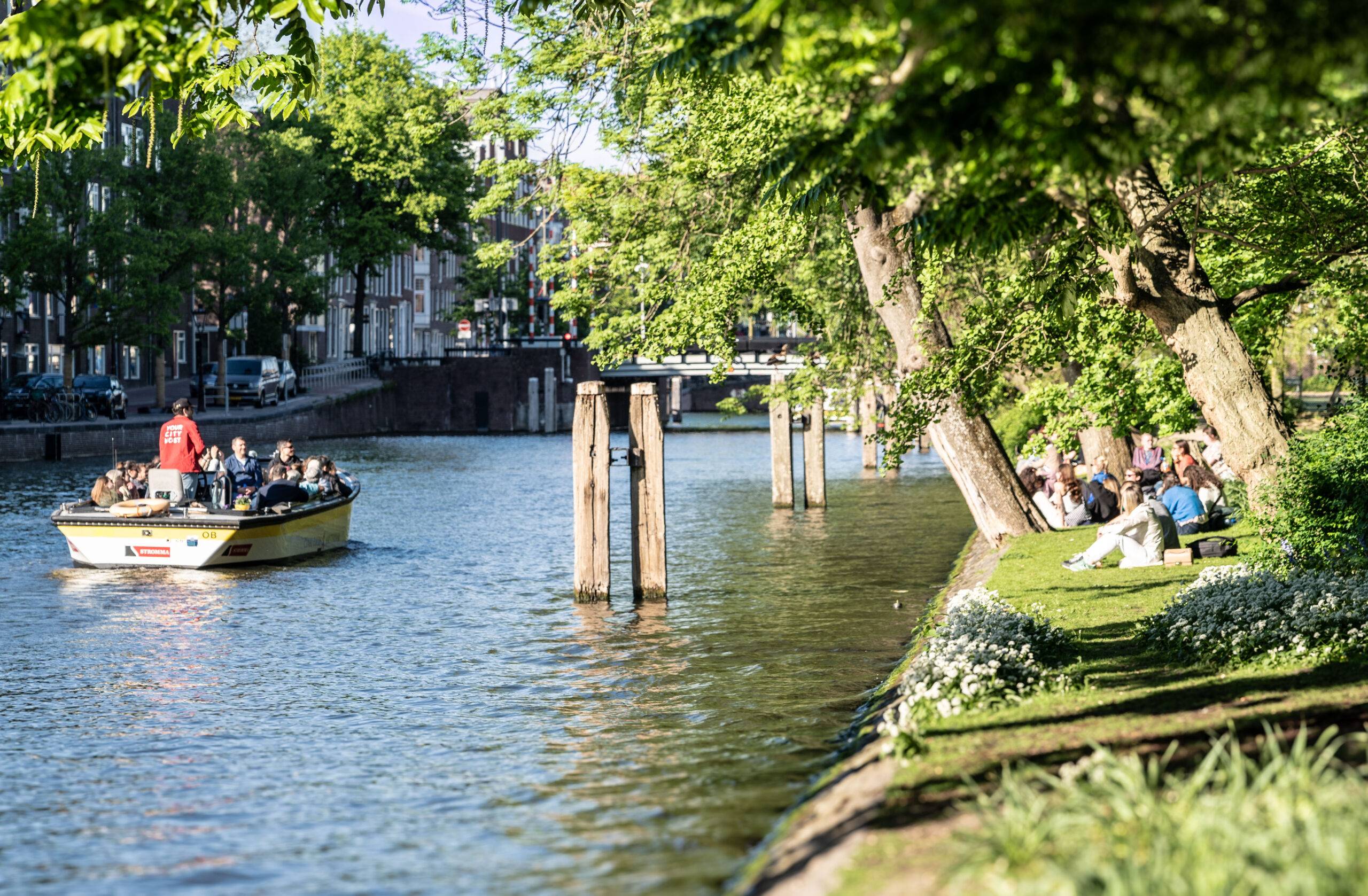 Menschen sitzen am Wasser im grünen Wertheimpark in Amsterdam, während ein Ausflugsboot vorbeifährt.