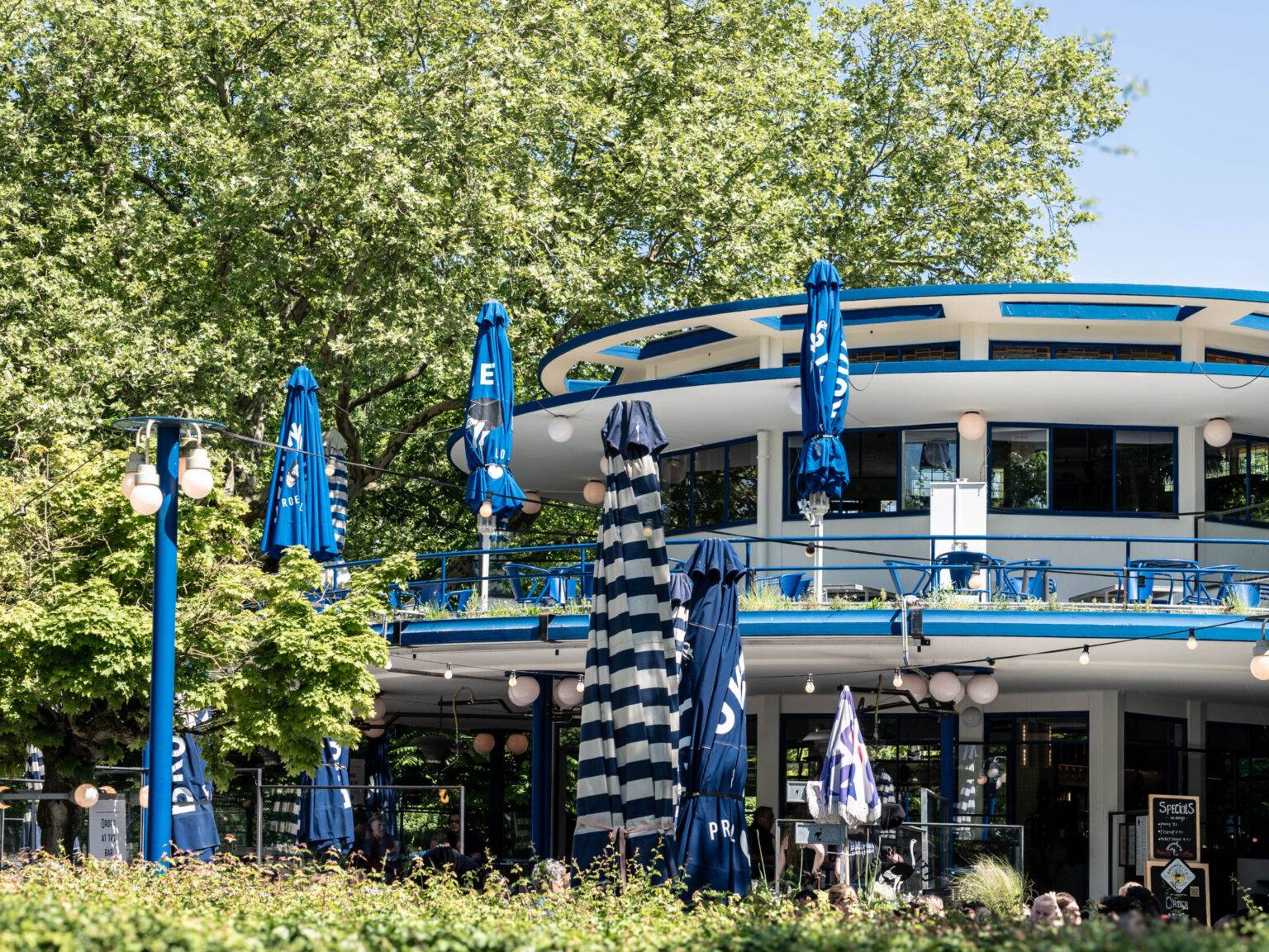 Das Blaue Teehuis im Vondelpark in Amsterdam, ein rundes modernes Gebäude im Bauhaus-Stil, umgeben von Bäumen und Besuchern.