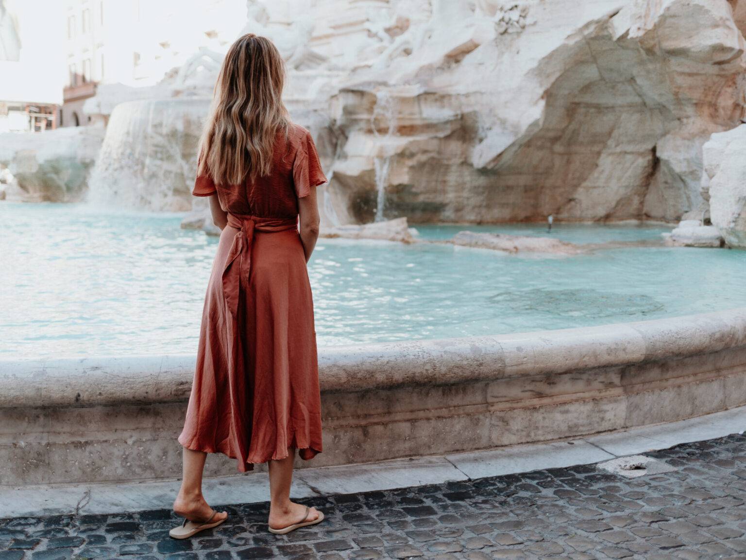 Frau im Sommerkleid steht am unteren Rand des Trevi-Brunnens in Rom und blickt zum türkisblauen Wasser und den barocken Figuren.