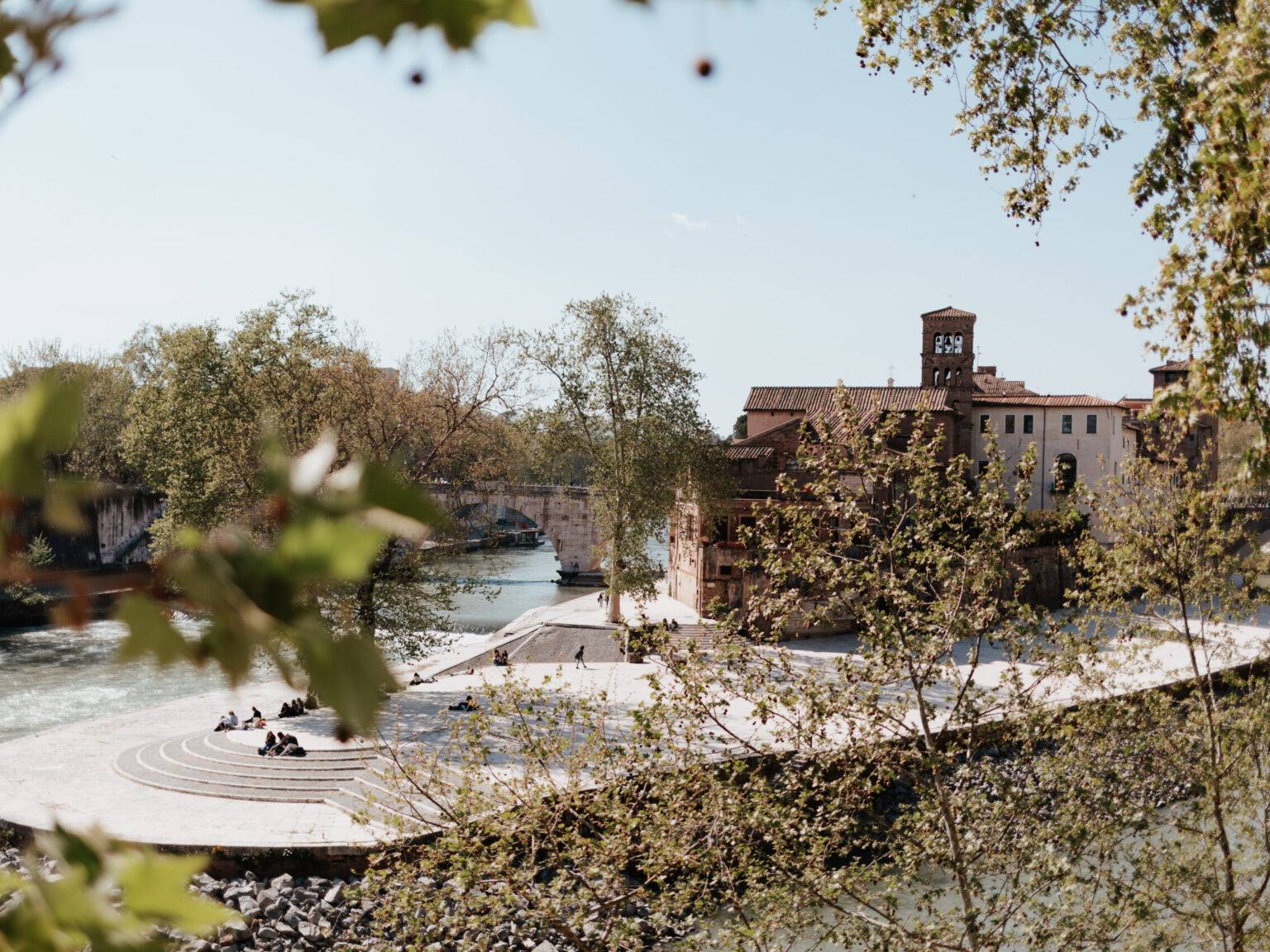 Die Tiberinsel in Rom mit Fluss, Bäumen, Spaziergängern und historischem Gebäude.
