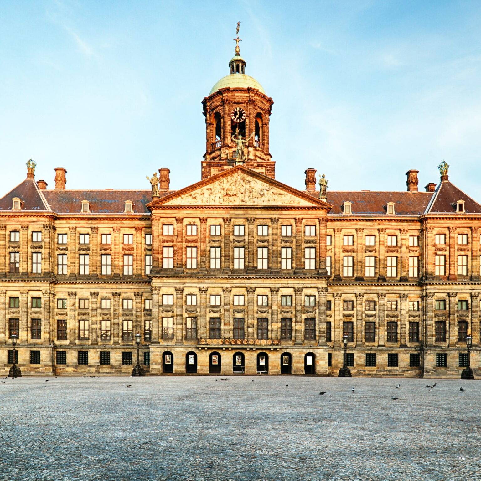 Die Fassade des Königspalasts in Amsterdam, einem großen, aus hellem Stein gemauertem Gebäude mit Uhrturm.