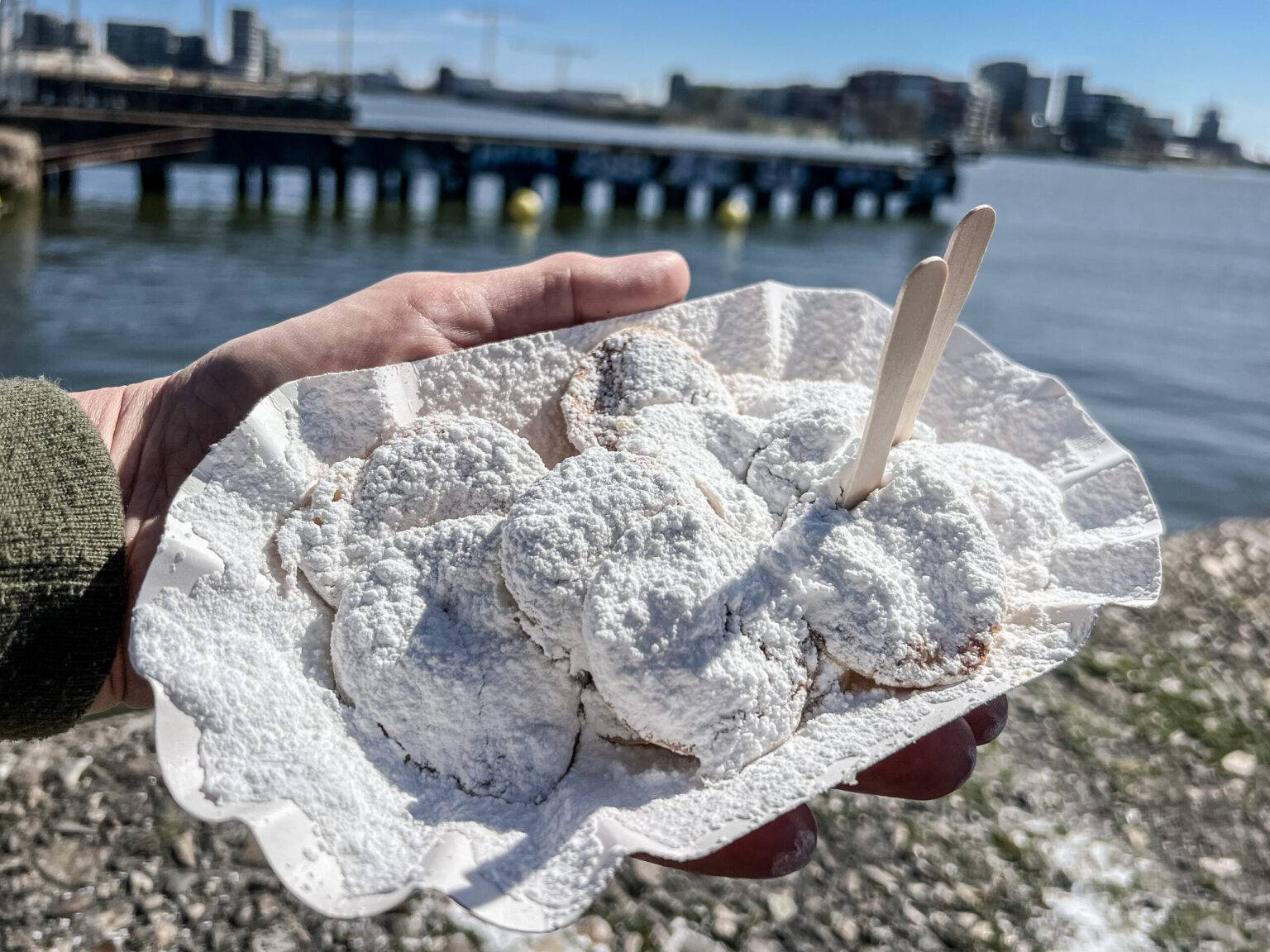 Eine Person hält einen Papierteller mit Poffertjes, die mit Puderzucker bestreut sind. Im Hintergrund ist Wasser und ein Steg.