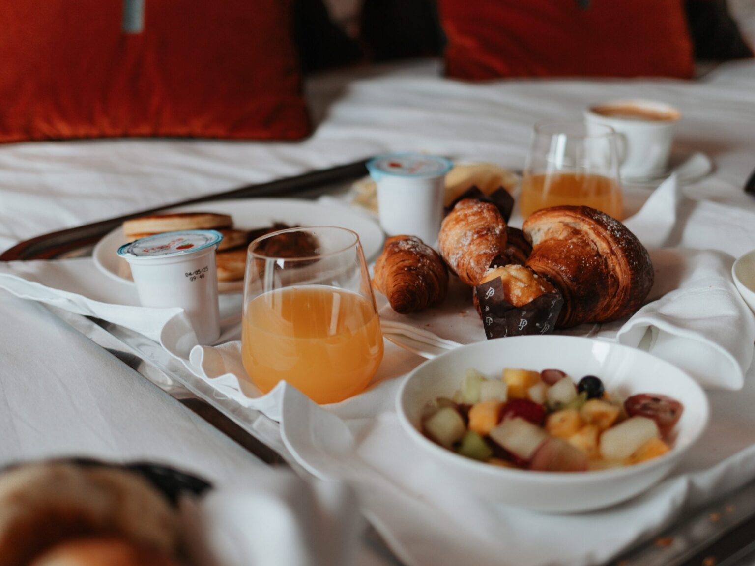 Ein Tablett mit geschnittenem Obst, Joghurt, süßen Backwaren und Orangensaft auf einem Bett in einem Hotelzimmer im The Tribune.