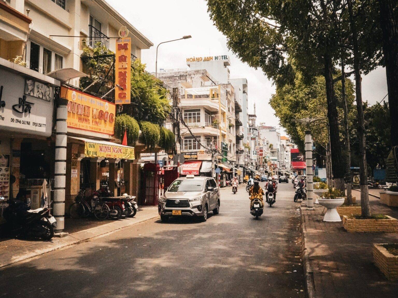 Eine Straße in Can Tho mit Rollern und einem Taxi gesäumt von Häusern und Bäumen.