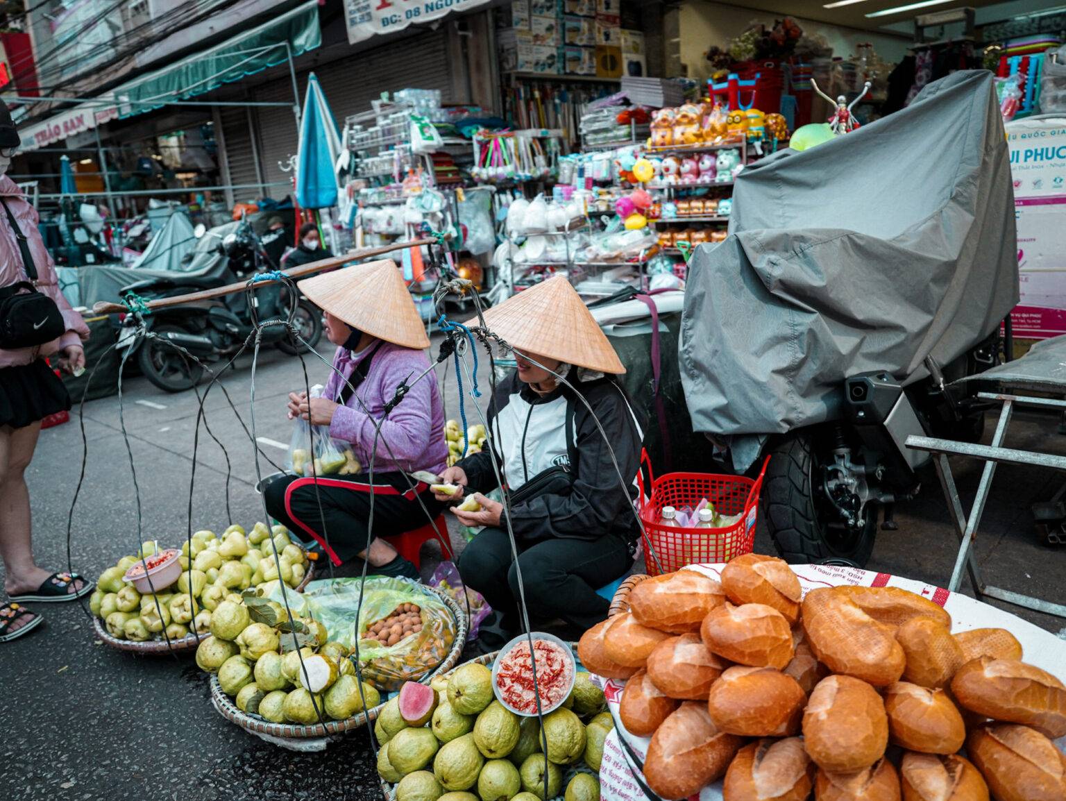 Auf der Straße auf dem Zentralmarkt von Da Lat sitzen zwei Händlerinnen mit Körben voller Obst.