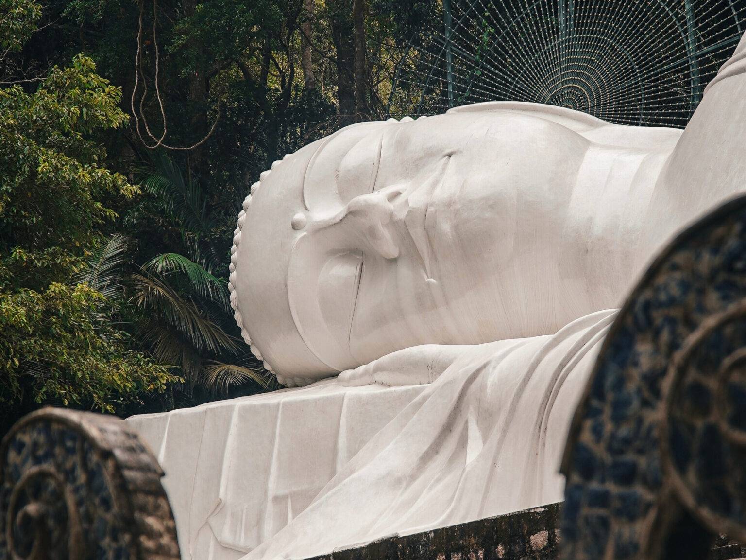 Ein gigantischer weißer liegender Buddha inmitten einer tropischen Landschaft.