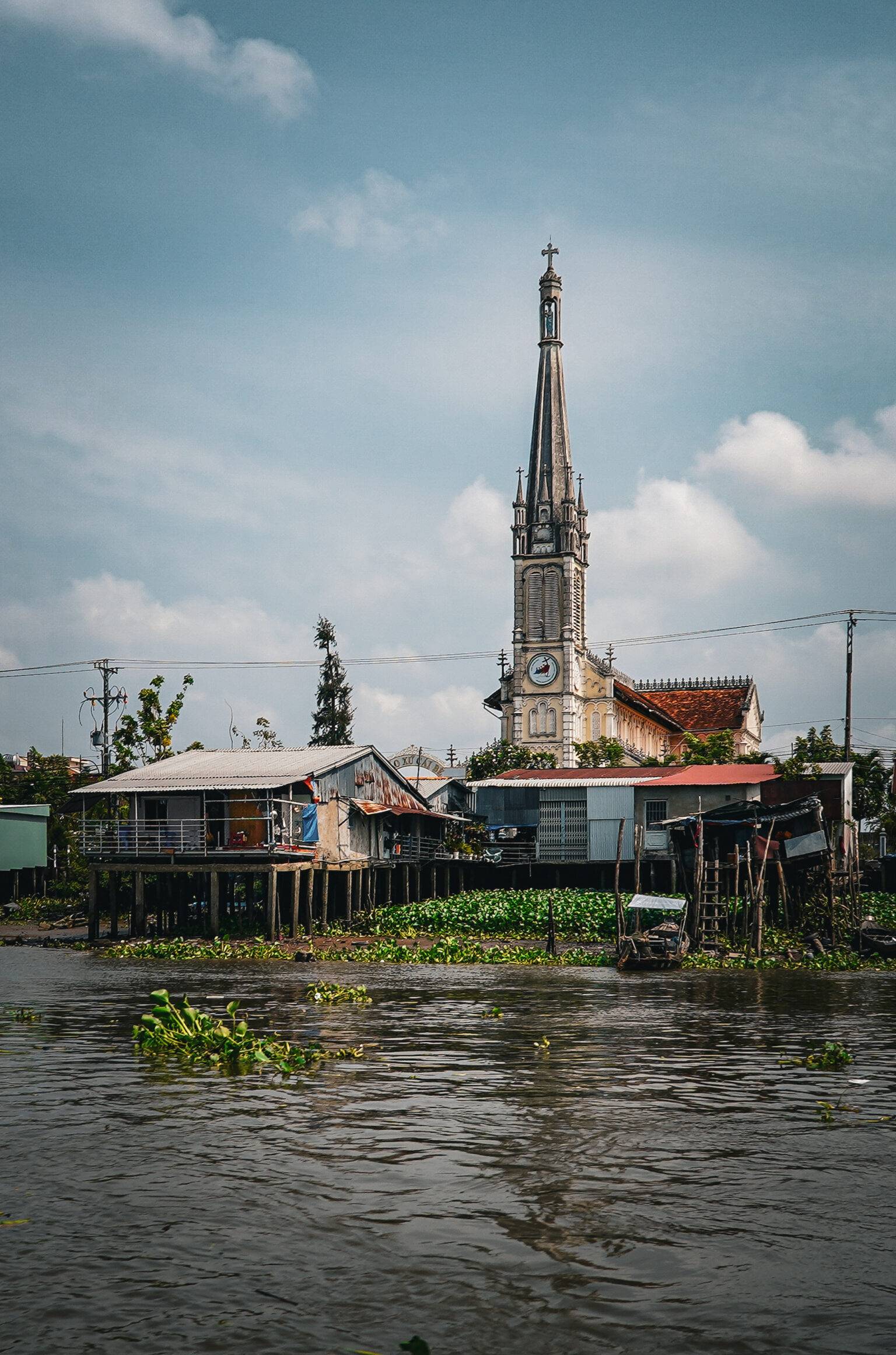 Die Kirche von Cai Be mit einem hohen spitzen Turm und dem Fluss im Vordergrund.