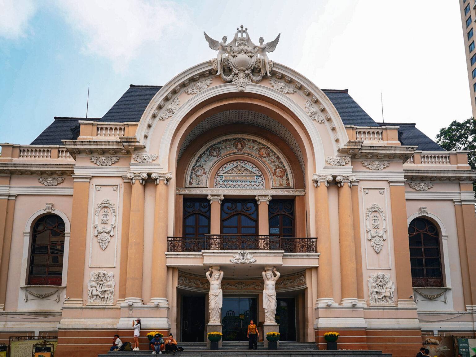 Die Fassade der Oper von Saigon mit einem großen zentralen Bogen, einem Balkon und vielen prunkvollen Verzierungen aus weißem Stein.