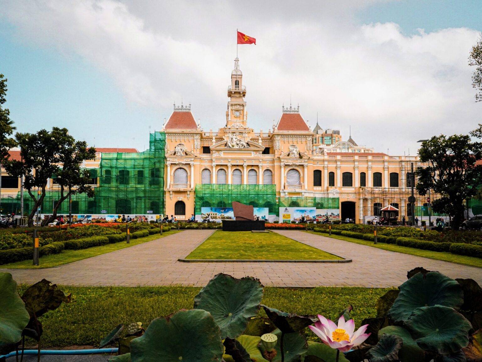 Die gelbe Fassade des Rathauses von Ho Chi Minh City mit einem Turm in der Mitte auf dem die Flagge von Vietnam weht. Das Gebäude ist teilweise mit einem Baugerüst verkleidet. Davor ist eine kleine, grüne Parkanlage.