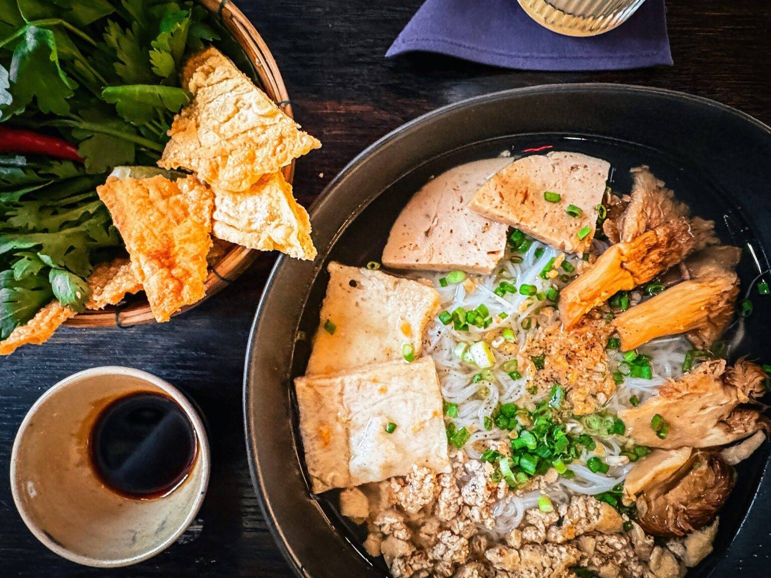 Auf einem Tisch steht eine Schale mit Nudelsuppe, eine Schale mit frischen Kräutern und ein Becher mit Sojasauce.