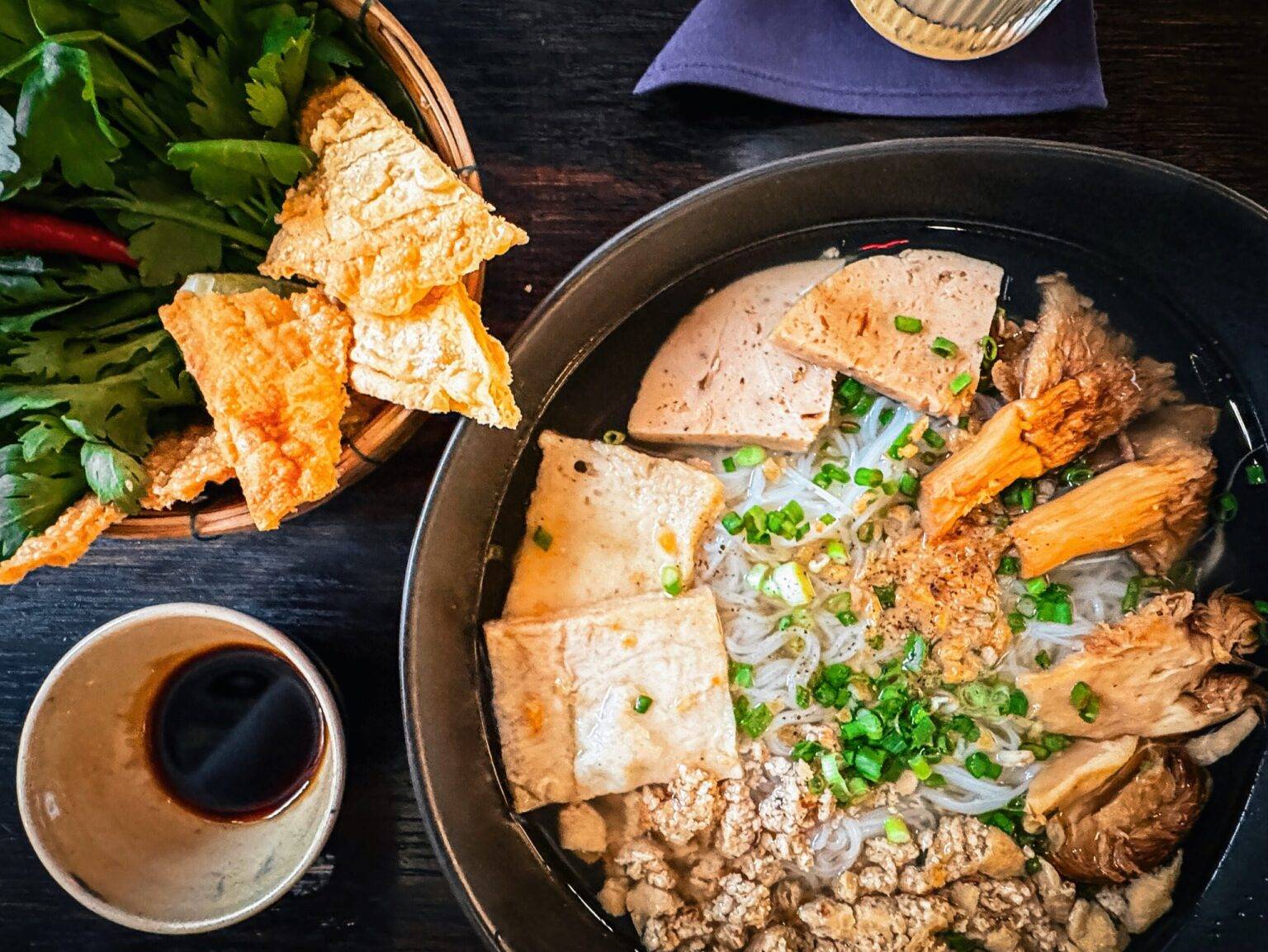 Auf einem Tisch steht eine Schale mit Nudelsuppe, eine Schale mit frischen Kräutern und ein Becher mit Sojasauce.