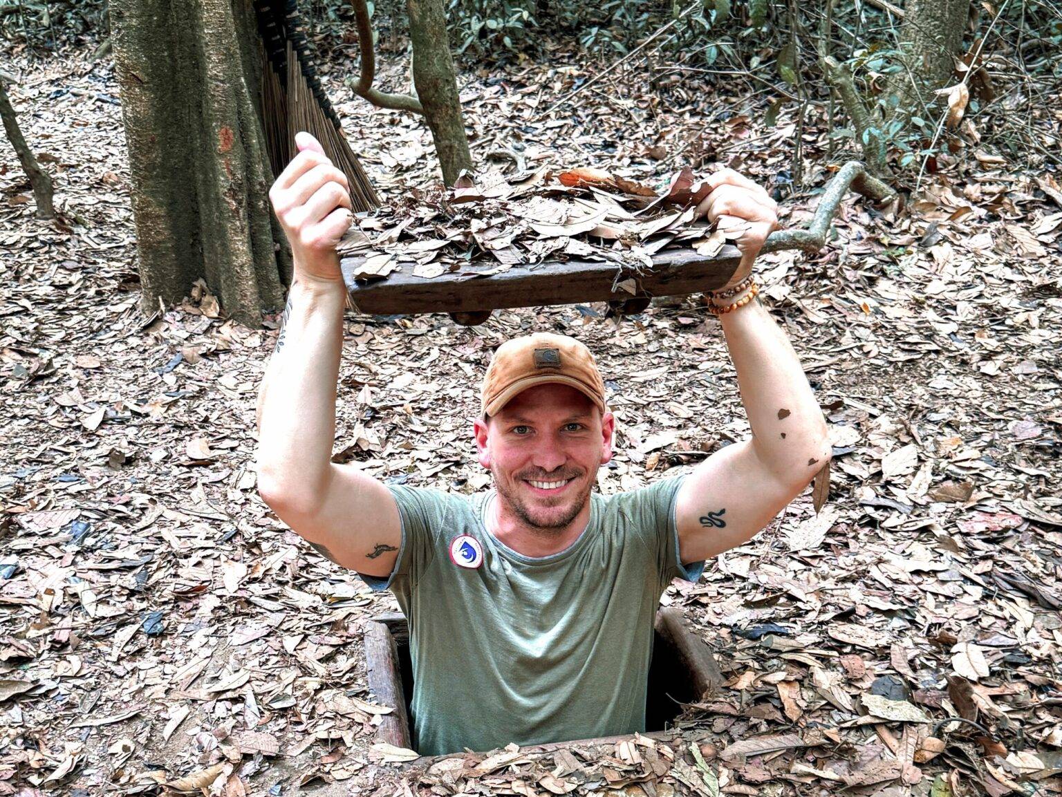 Daniel steht in der Öffnung zu einem Tunnel im Boden und hält den Deckel zur Tunnelöffnung über seinen Kopf uns lächelt in die Kamera. Der Boden im Wald ist mir Laub bedeckt.