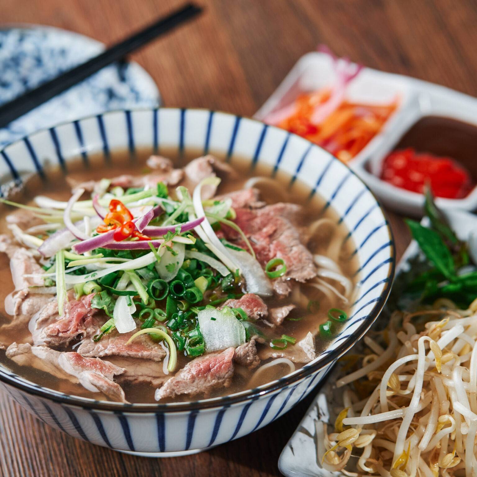 Eine Schale mit der Nudelsuppe Pho, mit Fleisch und frischen Kräutern. Daneben ein Teller mit Sprosse und kleine Schälchen mit Saucen.
