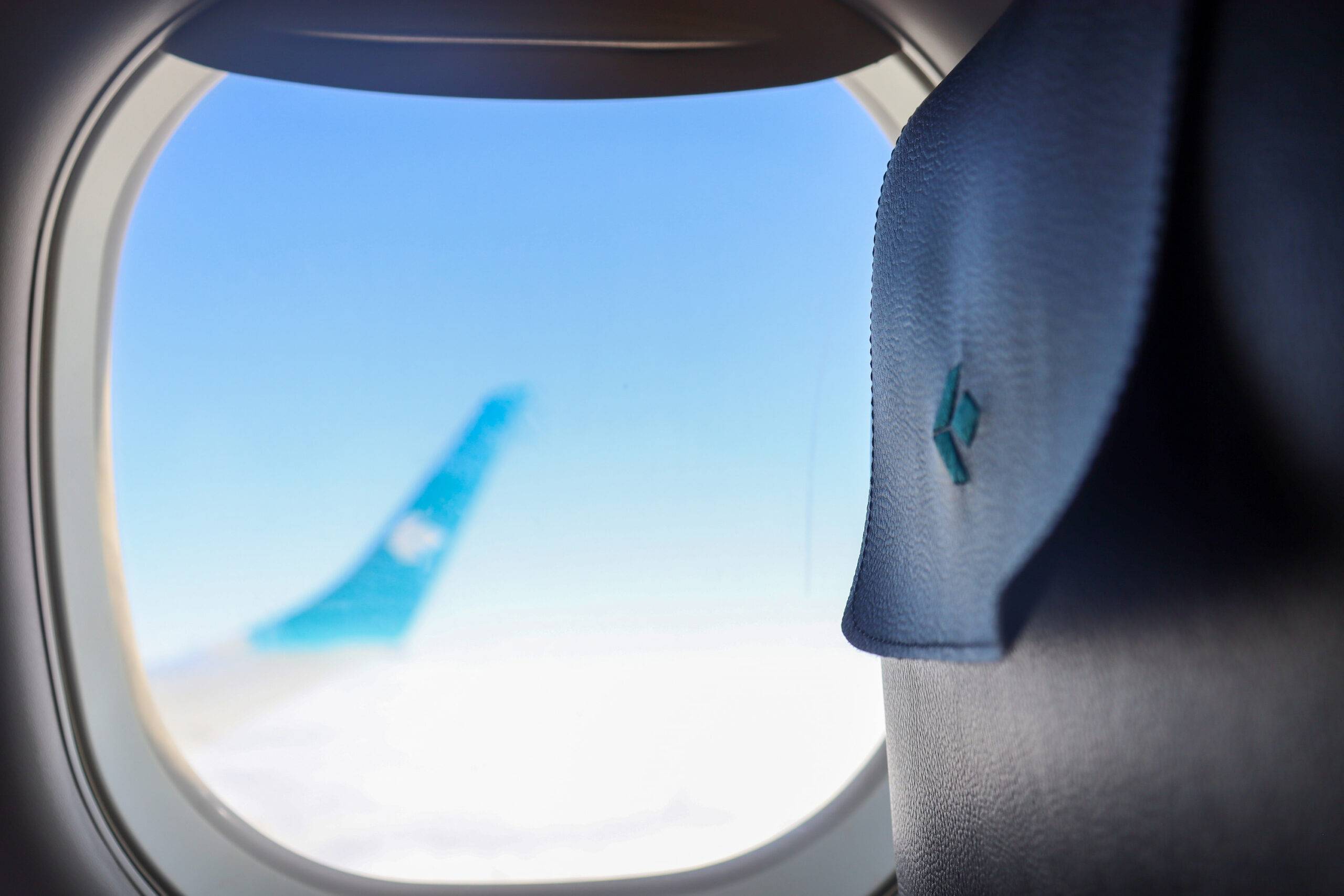 Das Kopfteil eines Sitzes in einem Flugzeug von Air Dolomiti neben dem Flugzeugfenster. Verschwommen im Hintergrund die Tragfläche über den Wolken.