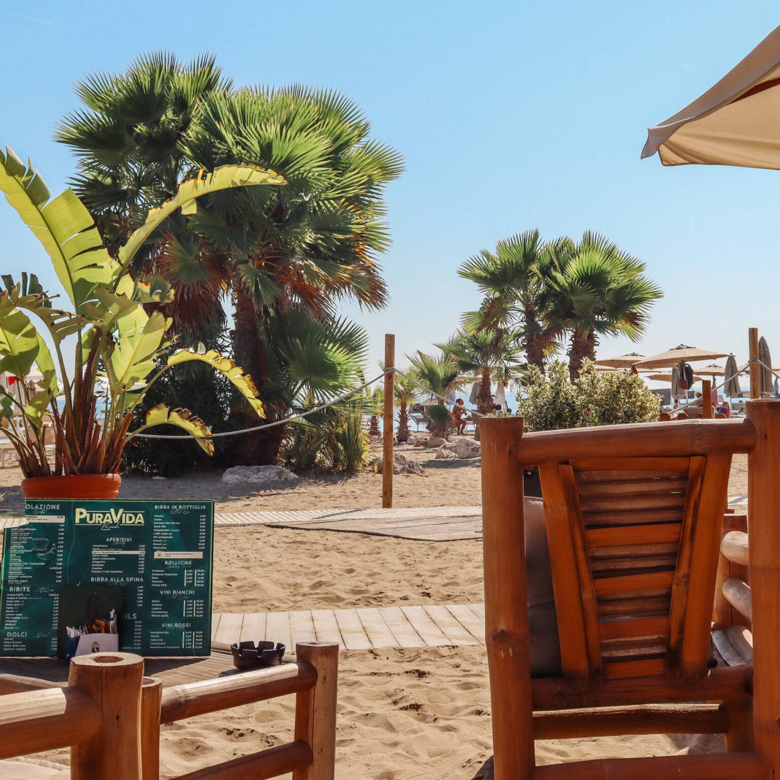 Die Strandbar Pura Vida Beach mit Bambusstühlen im Sand und Blick auf Palmen und Sonnenschirme.