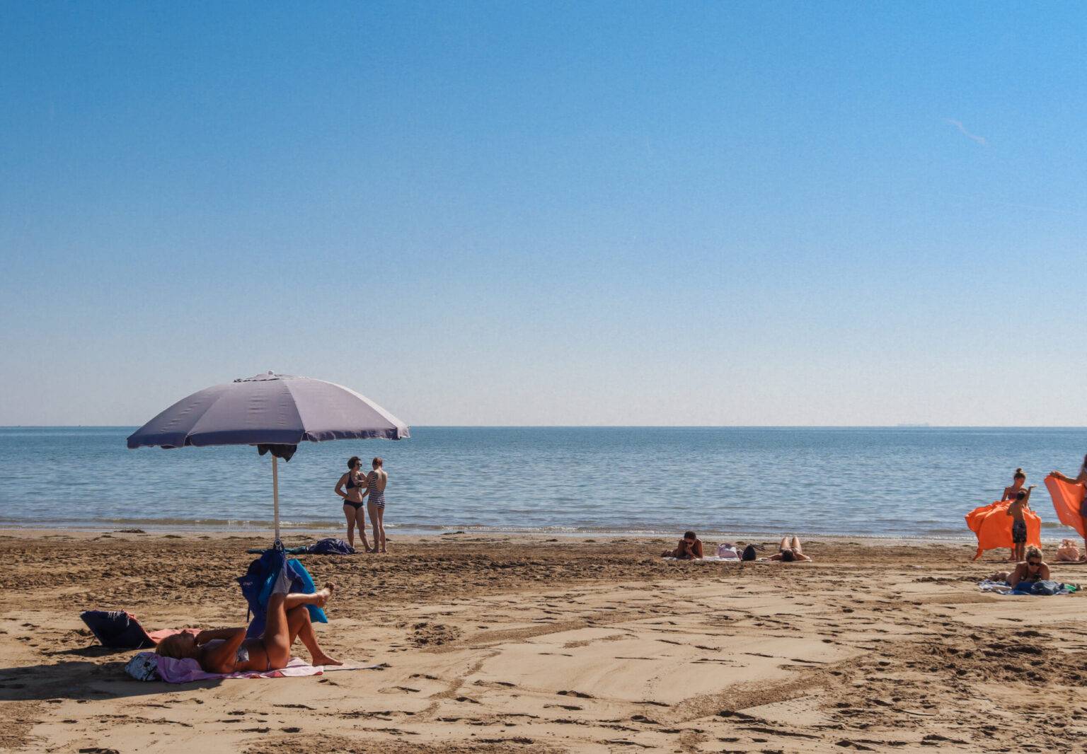 Der lange Sandstrand und das Meer beim Strandbad Bluemoon. Vorne links sitzt eine Person unter einem Sonnenschirm.